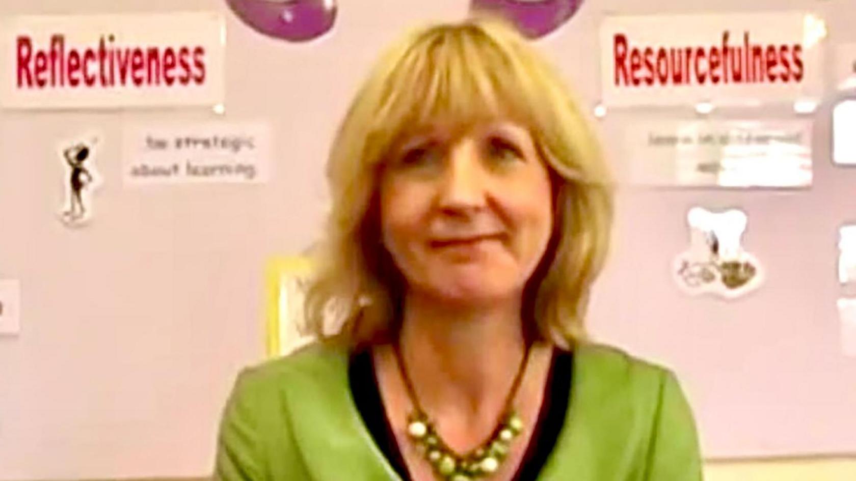 Christine Baker is seen in front of a school noticeboard showing a display about learning strategies. She has blonde hair with a fringe and is wearing a green jacket with a black top and a necklace of green beads.