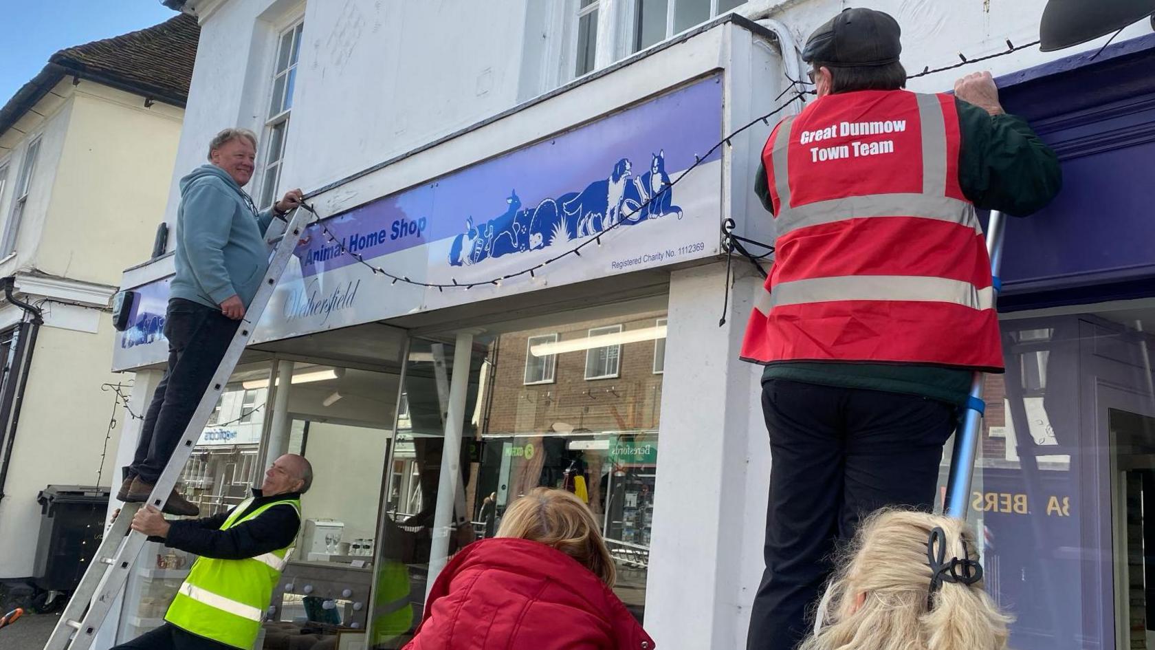 People use ladders to fix a long line of Christmas lights to a blue and white shopfront. A man is up a ladder wearing a blue hooded top. A man wearing a high vis yellow jacket is supporting the ladder. Another person, in a red vest, is on another ladder.
