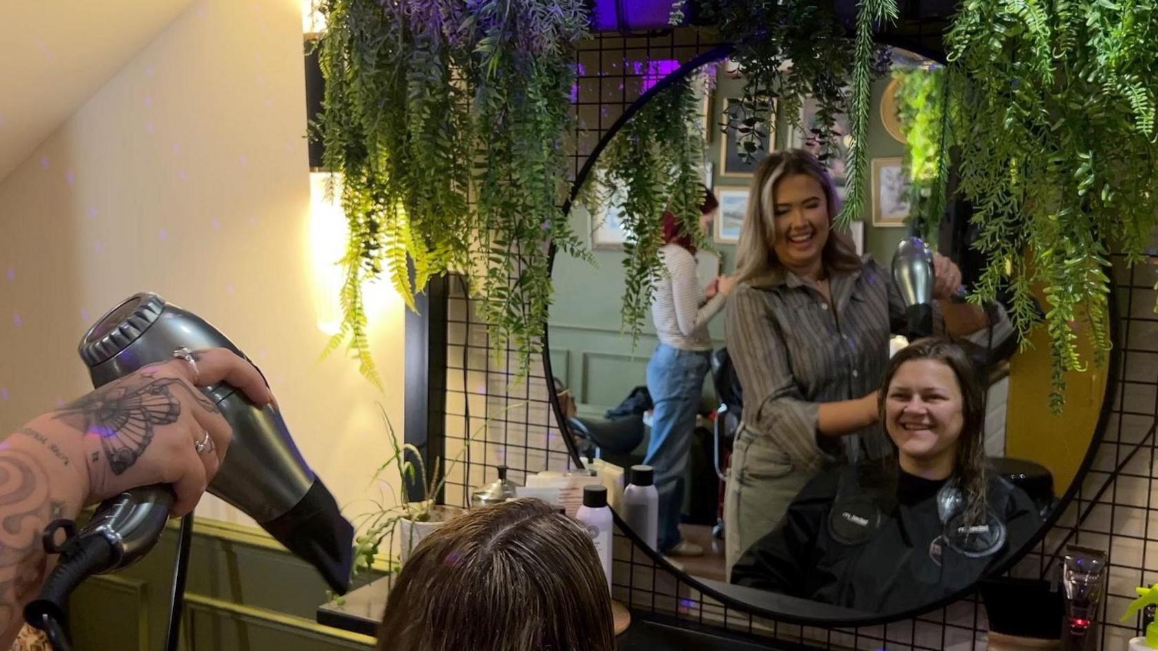 The imagine shows a women having her hair blow-dried by another woman in a hair salon. The picture has been taken from behind the client and the women's' reflection can be seen in a round mirror in front of them. The hairdresser's hand can be seen in the bottom left corner of the image holding a hairdryer.