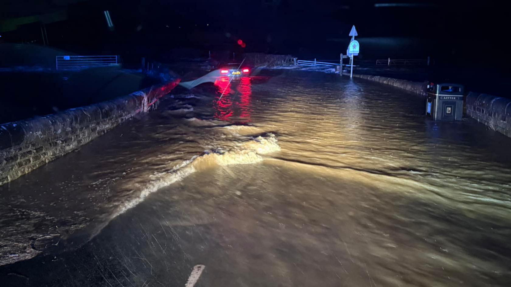 The rear brake lights of a car can be seen on a flooded road, which is in darkness but illuminated by the lights from a fire and rescue vehicle behind the camera.