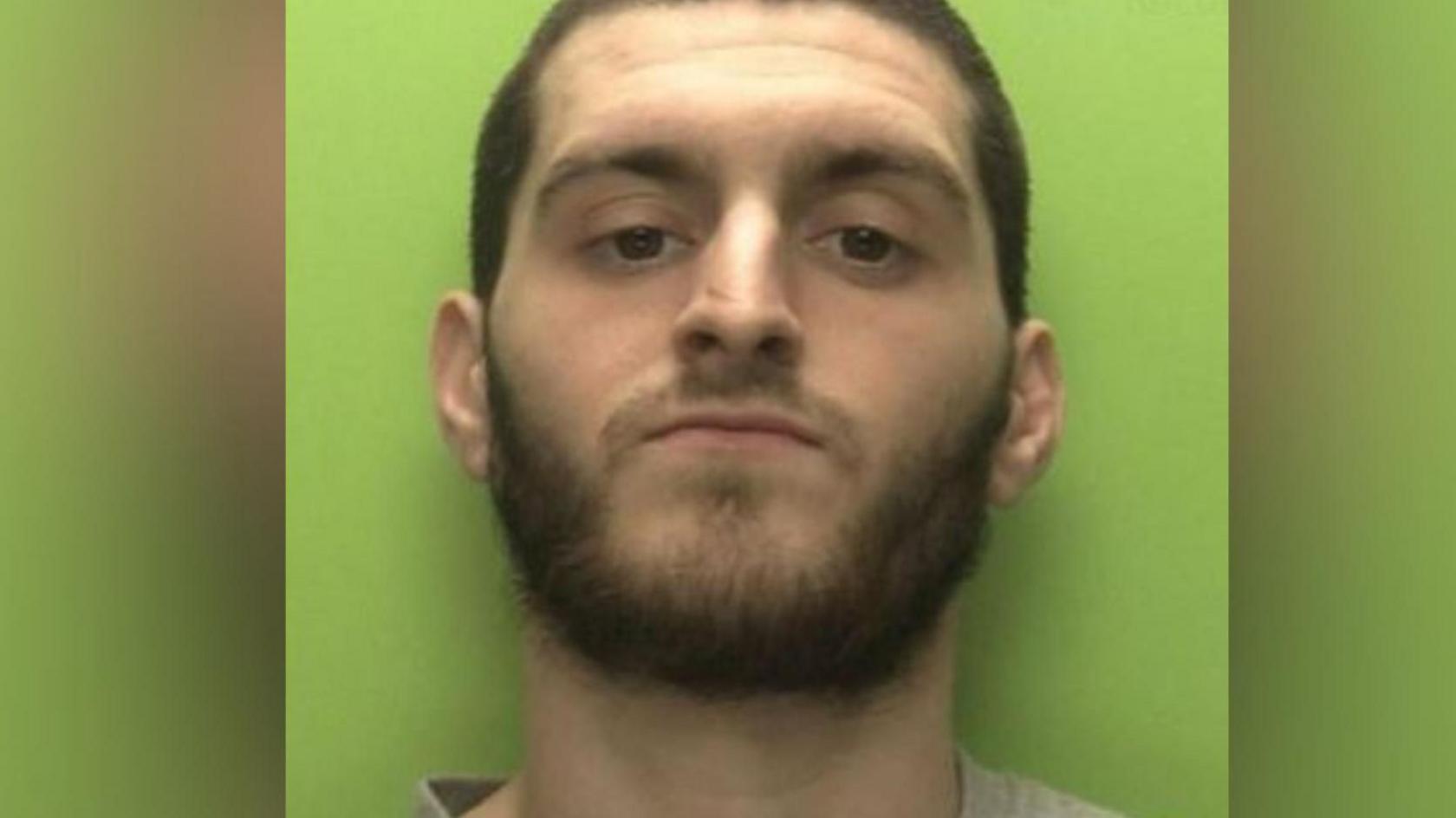 A mugshot of Connor Page, a 28-year-old white man with short brown hair all one length and a short beard. He is standing in front of a lime green backdrop.
