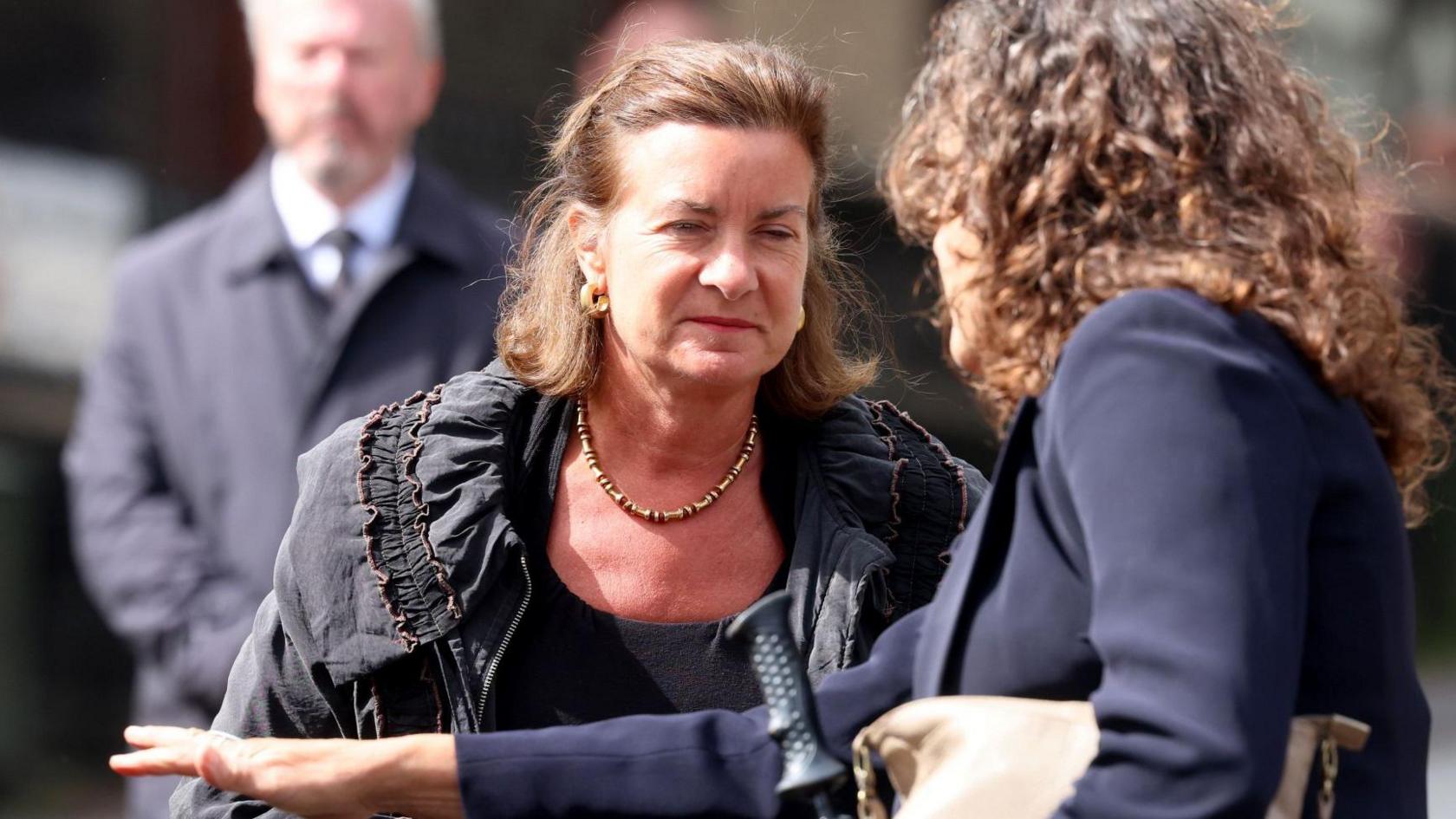 Eluned Morgan stood holding a bag next to a colleague, whose stood with the back of her head to the camera. Eluned is squinting and is wearing a black top and black jacket.