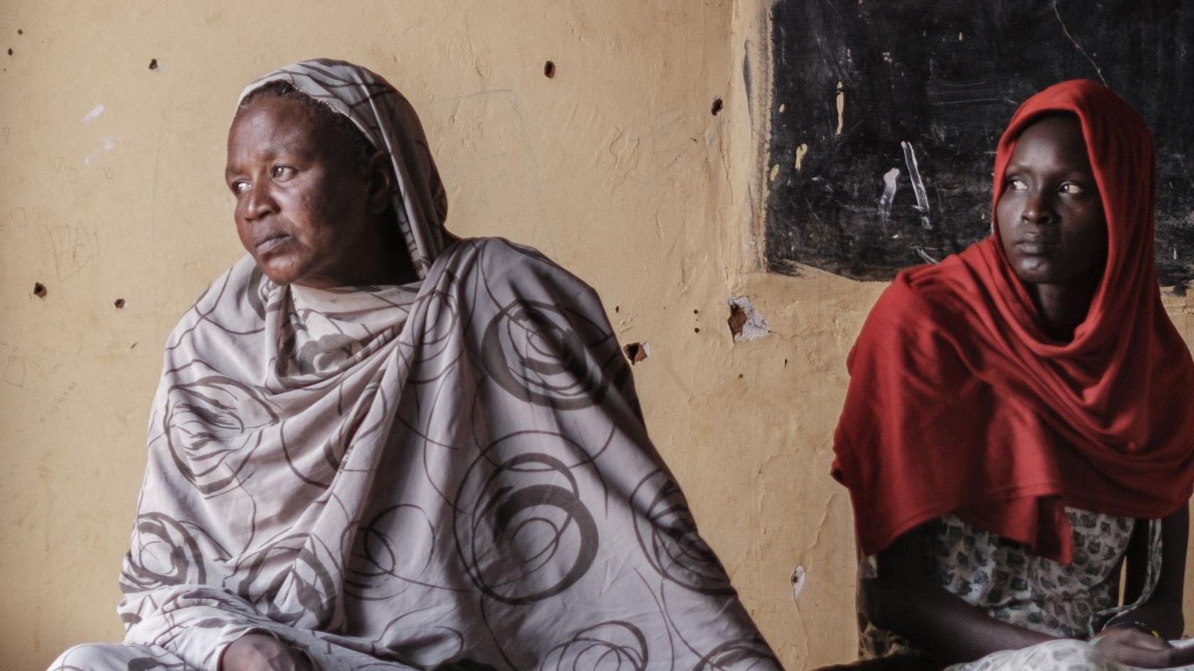 Two Sudanese women looking sad, sitting in a classroom at a school sheltering Internally Displaced People (IDPs) in the city of Omdurman, Sudan. A wall with a blackboard in the background.