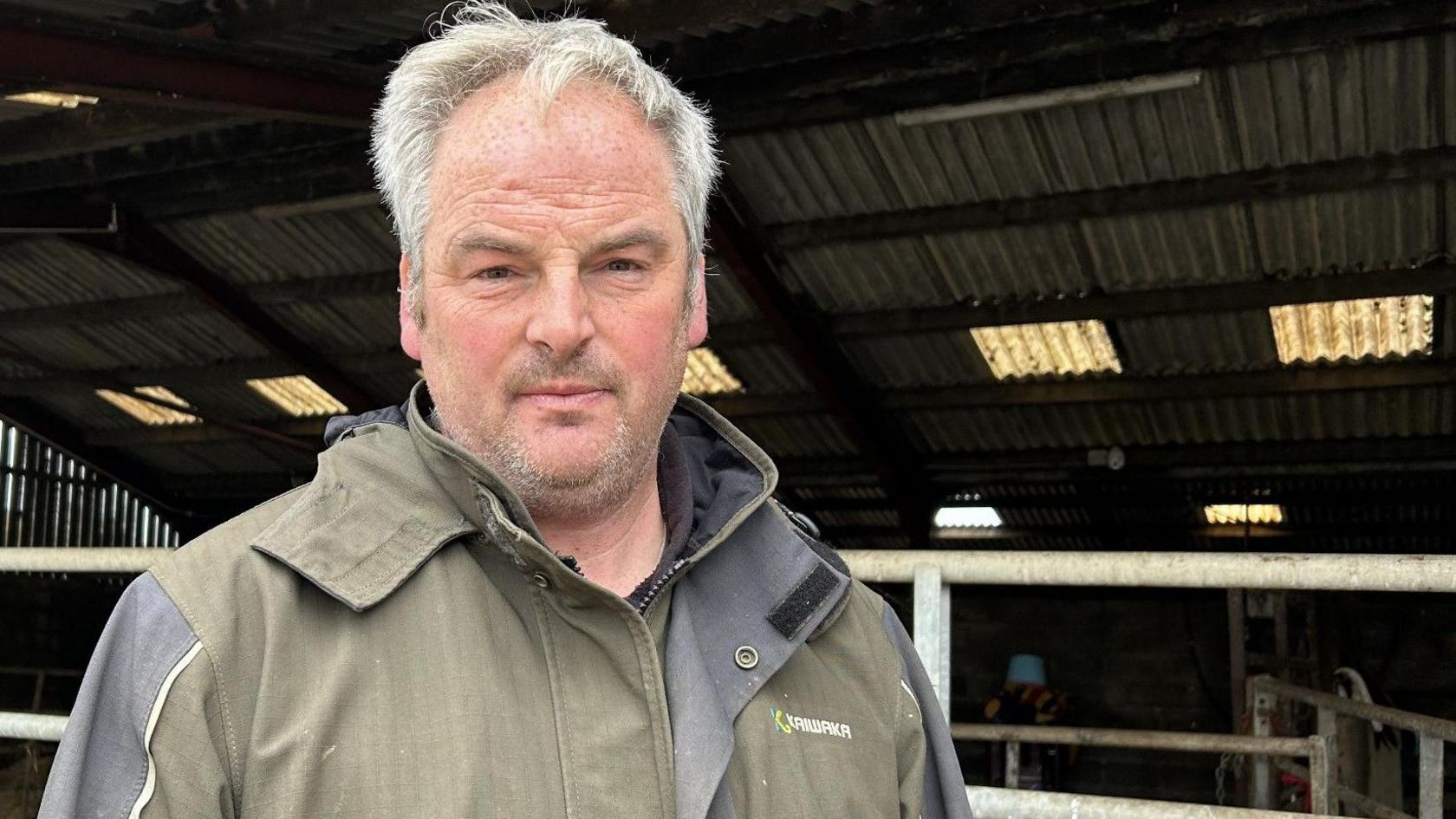 Mark Davies, wearing a green coat, looks at the camera while standing near a farm shed