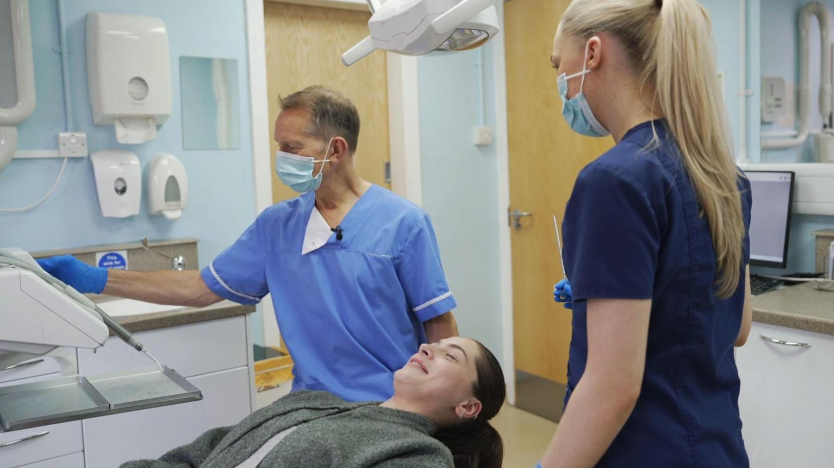 Glasgow dentist David McColl with a patient undergoing a check-up - she is lying down and smiling. He is in his medical scrubs and checking a machine. A dental nurse is standing next to the patient, with her back to the camera.   