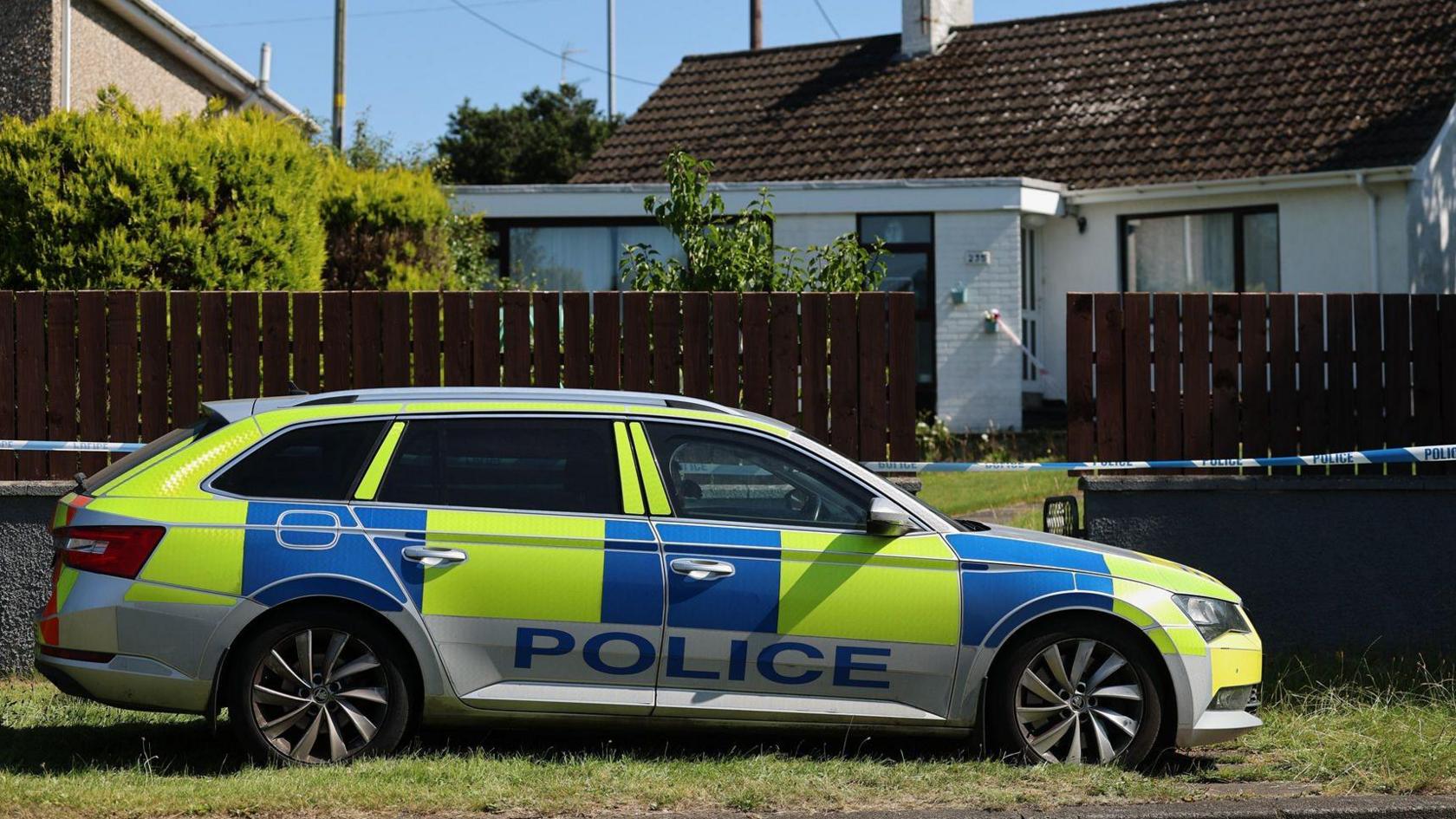 A yellow, blue and white police car outside the white bugalow with police tape visible 