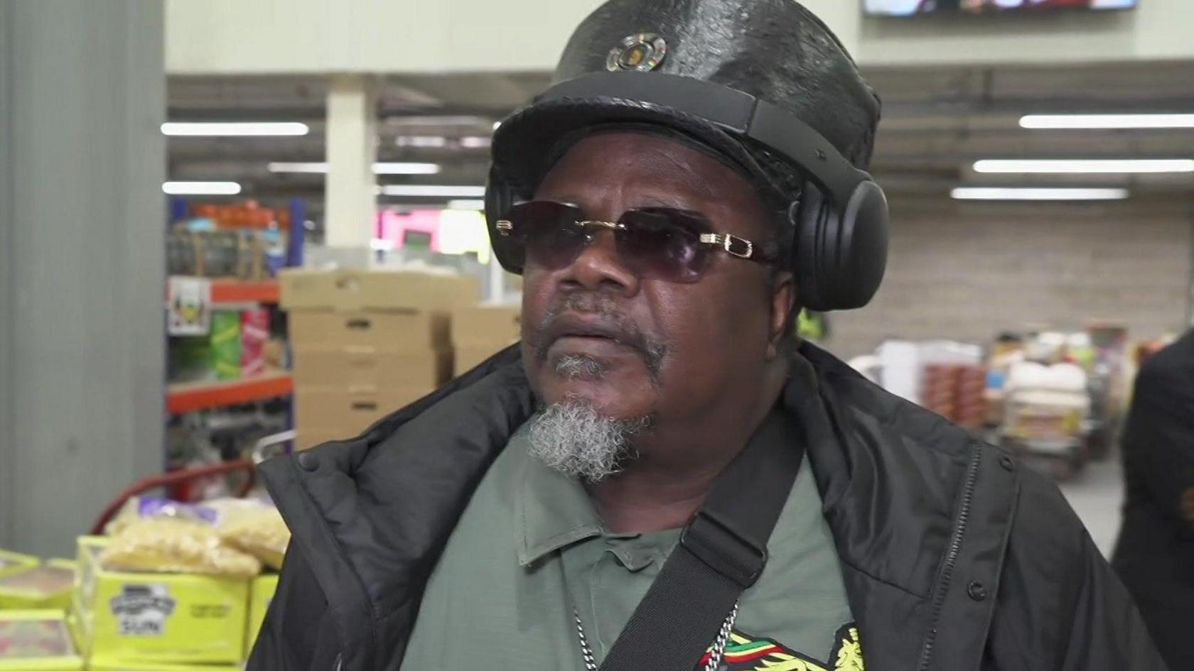 Reggae singer Luciano in front of the same grocery shop, wearing a leather hat, headphones and black sun glasses. Underneath his black jacket, he is wearing a khaki shirt and an over-the-sholder bag strap can also be seen