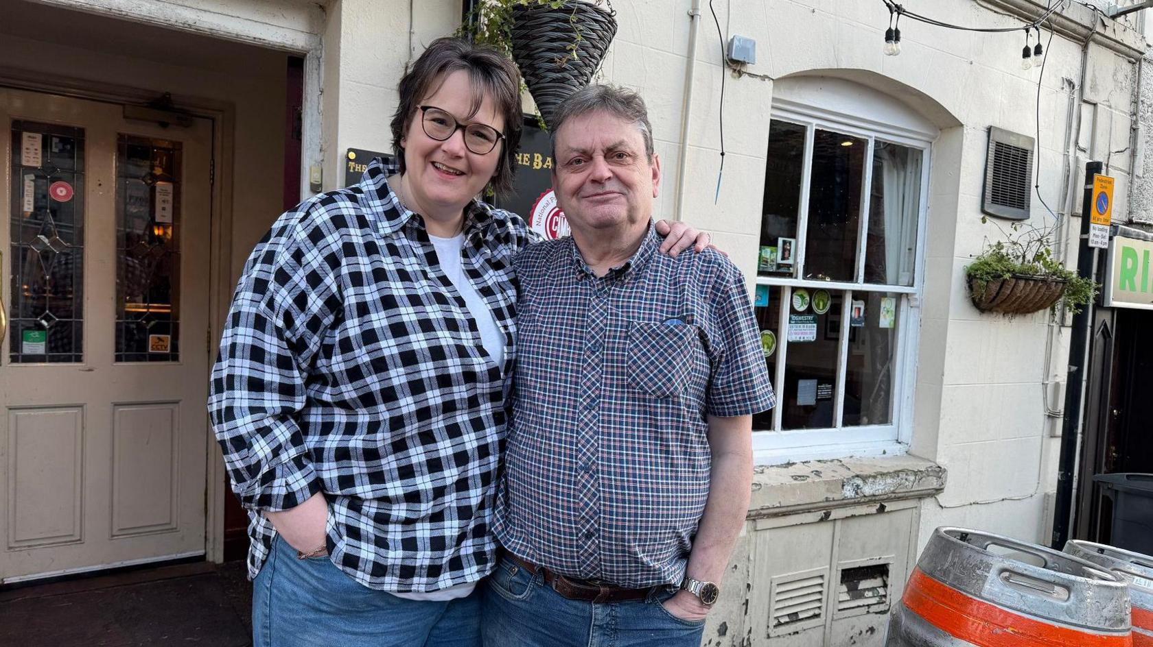 A man and a woman are standing outside a pub with cream white walls. The man has grey hair and is wearing a checked shirt. The woman has short broken hair and is wearing glasses. They have their arms around each other