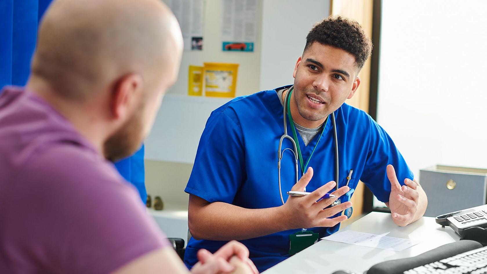 A young male doctor is talking to a male patient in a doctors office. We can see the doctors face but only the back of the patients head. The doctor is in blue scrubs and the patient is wearing a purple T -shirt