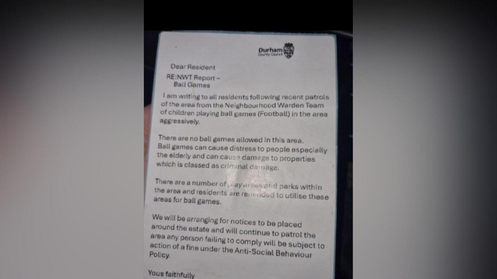A hand holding a laminated fake note, claiming to be from Durham County Council Neighbourhood Wardens - North team. The letter states it was sent over children playing football aggressively and claims no ball games are allowed in the area. It adds ball games can cause damage to property and distress. It ends with a claim that more patrols will be carried out and notices will be placed in the area. It says those failing to comply will be fined. Durham County Council's logo is printed in the top right corner.