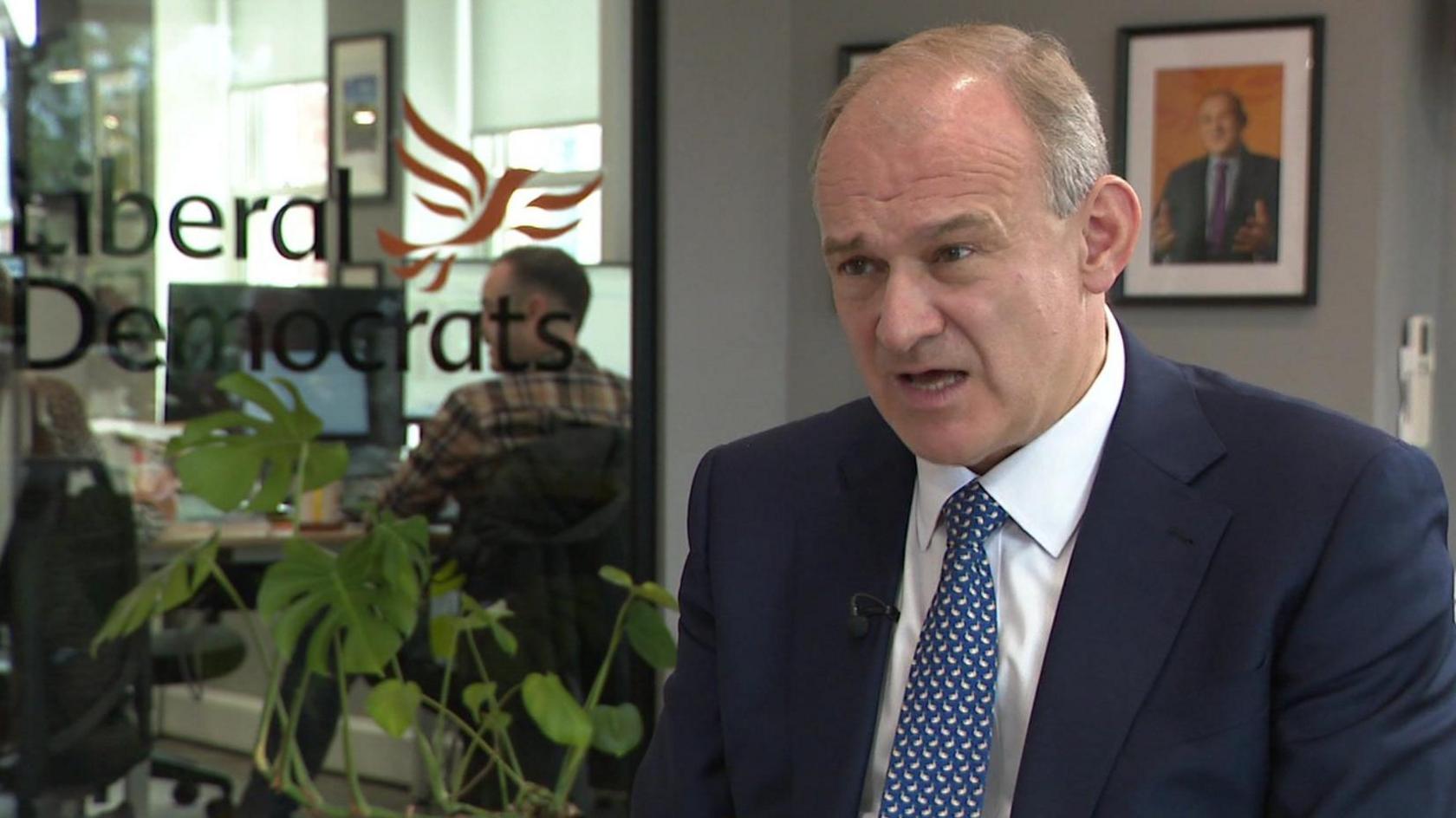 Sir Ed Davey being interviewed in Lib Dem HQ, wearing a blue suit and tie with people working in the background behind glass with "Liberal Democrats" and the party logo emblazoned on it 