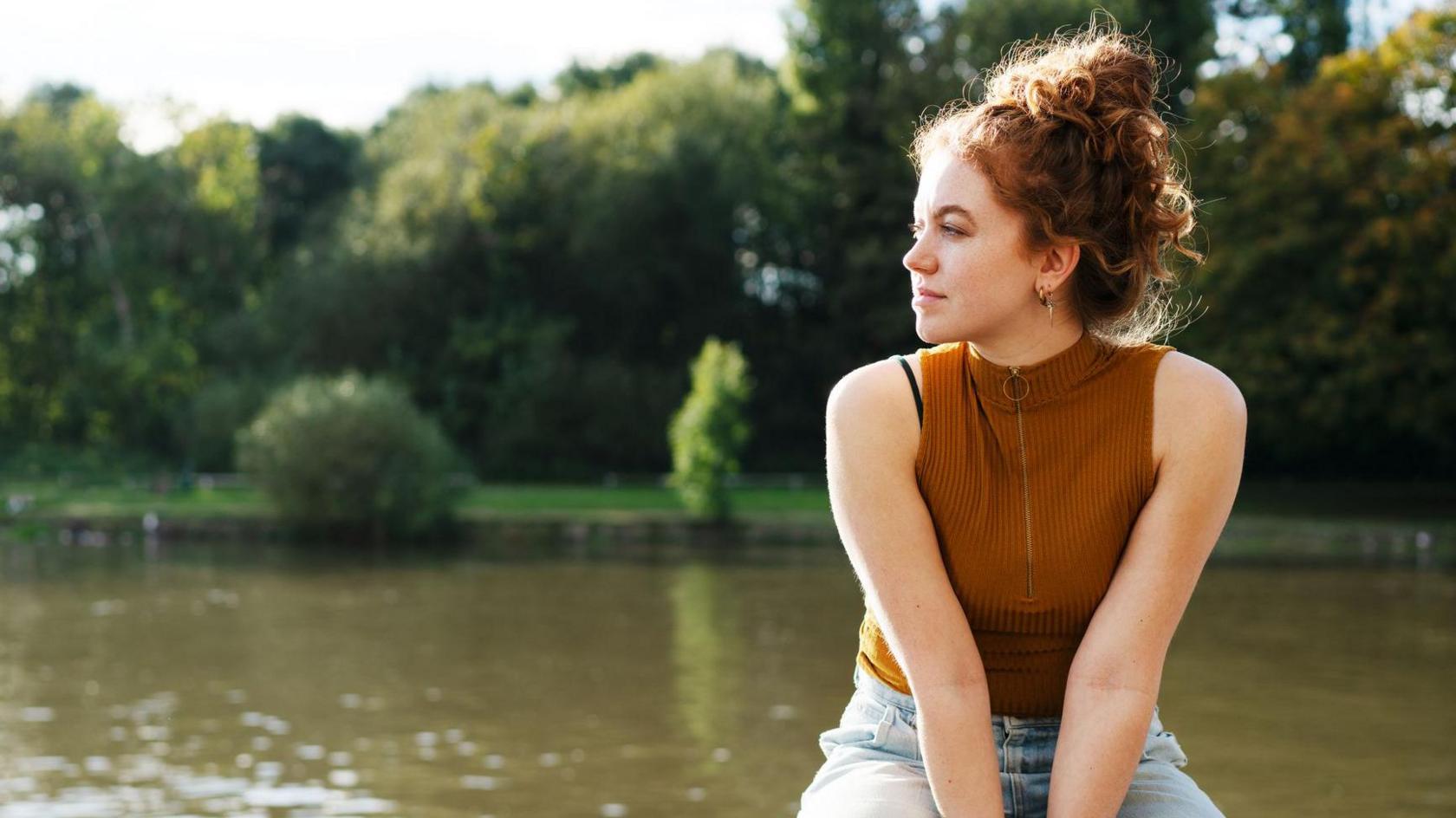 A woman wearing a brown top and blue jeans with ginger hair tied up, sitting on a ledge with a lake in the background. 