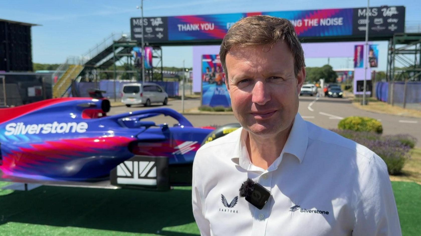 A head and shoulders shot of Stuart Pringle, outside at Silverstone. He has short, light brown hair and is wearing a white open-neck shirt, with a small black microphone attached to it. Behind him is a purple and red car with Silverstone written on it in white letters and beyond that a road with a bridge over it.