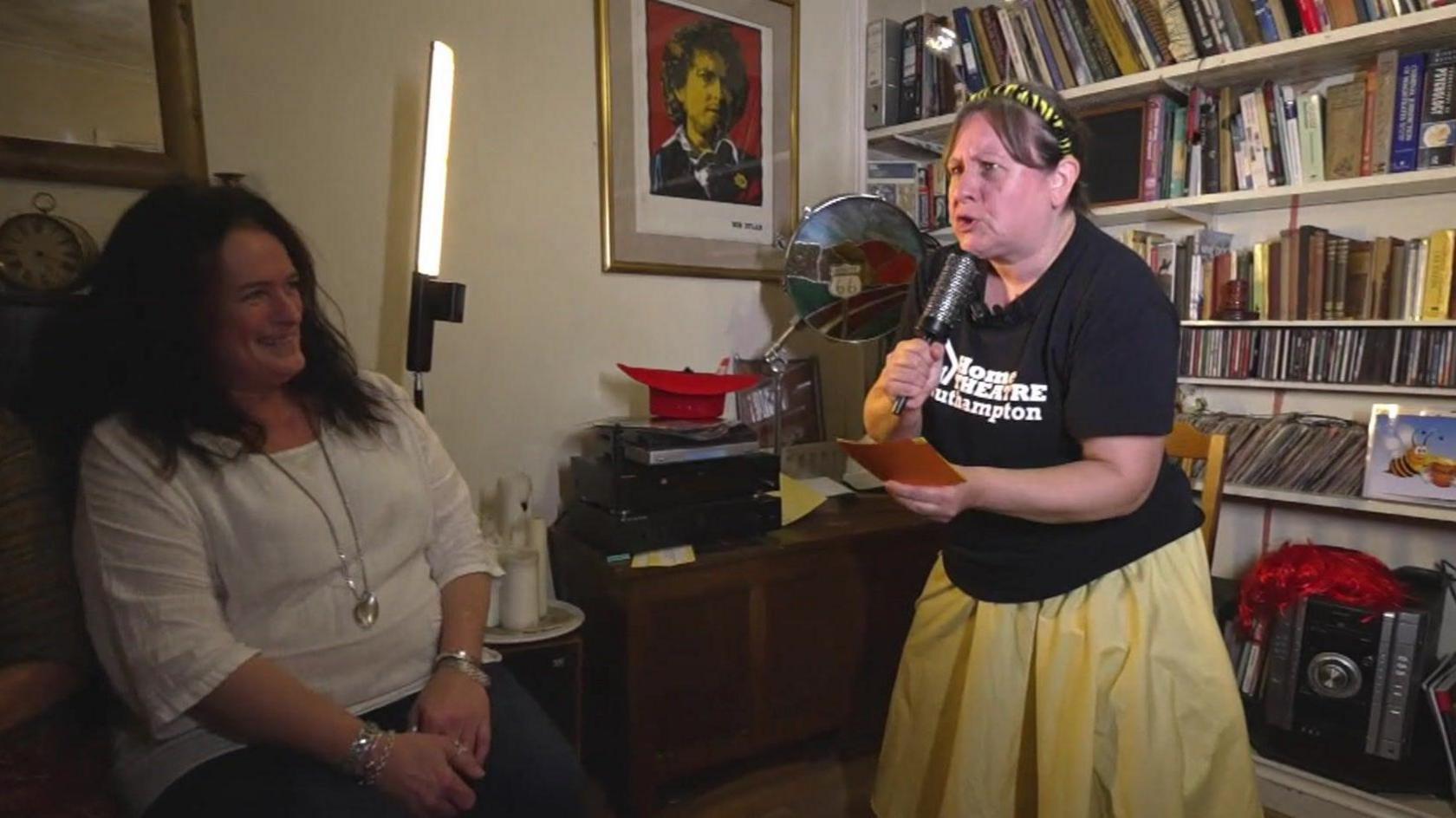 An actress, lit by a portable light stand, performs in a darkened living room. She is speaking into a pretend microphone, as if pretending to be a news reporter. A woman in the audience is laughing. Behind the actress are bookshelves and other home furniture.