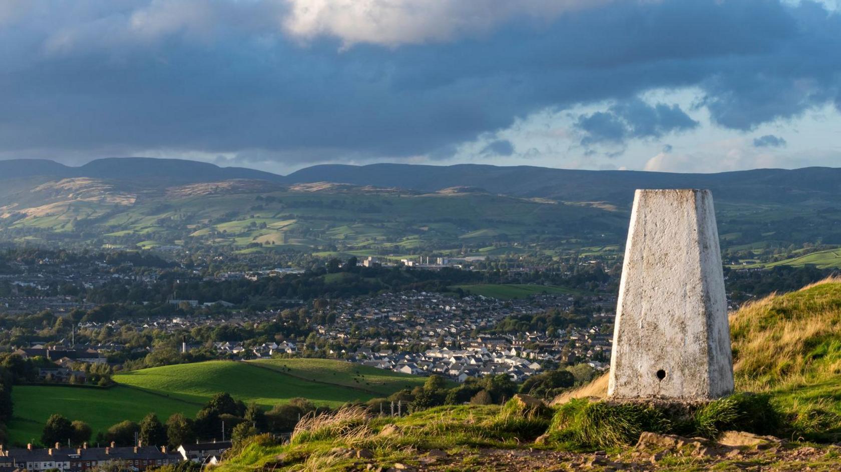 Trig pillar painted white can be seen sitting on a hilltop overlooking a town