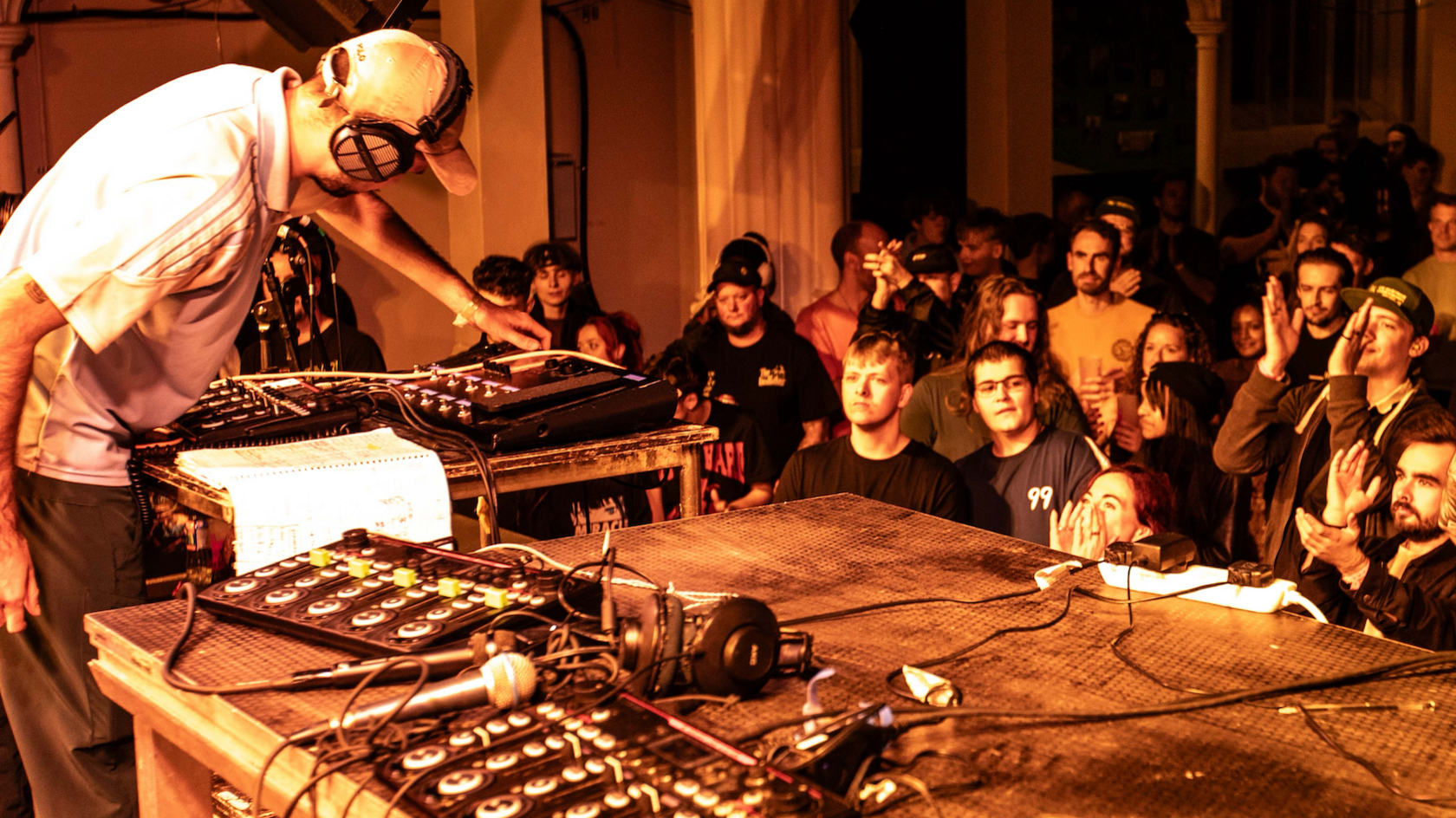 A performer with a DJ deck. The DJ is wearing a white cap with black headphones over it, and a white t-shirt. The picture includes all of the sound equipment on the stage. 