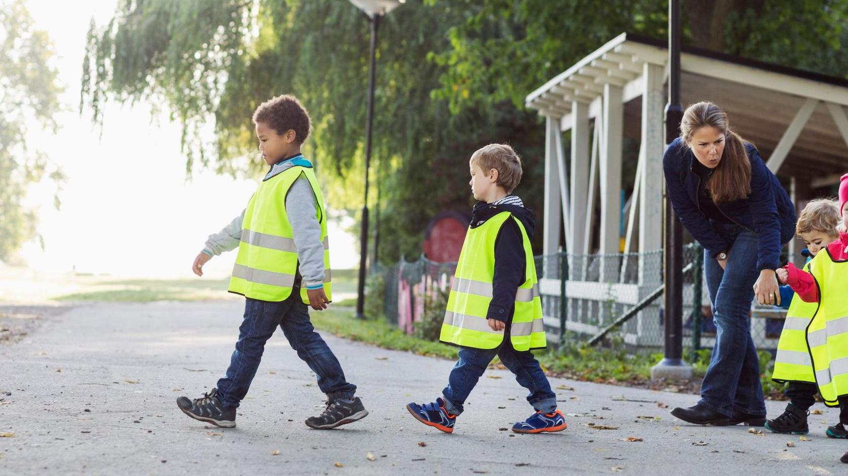 Two small children wearing oversized hi-vis jackets crossing a road. A teacher is waiting with smaller children at the side of the road 