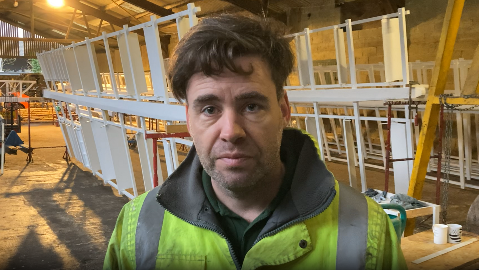 Timothy Whiting looks at the camera in a warehouse. White windmill sails rest behind him in the warehouse. He has brown hair that overhangs on his forehead and he is wearing a florescent green coat. 