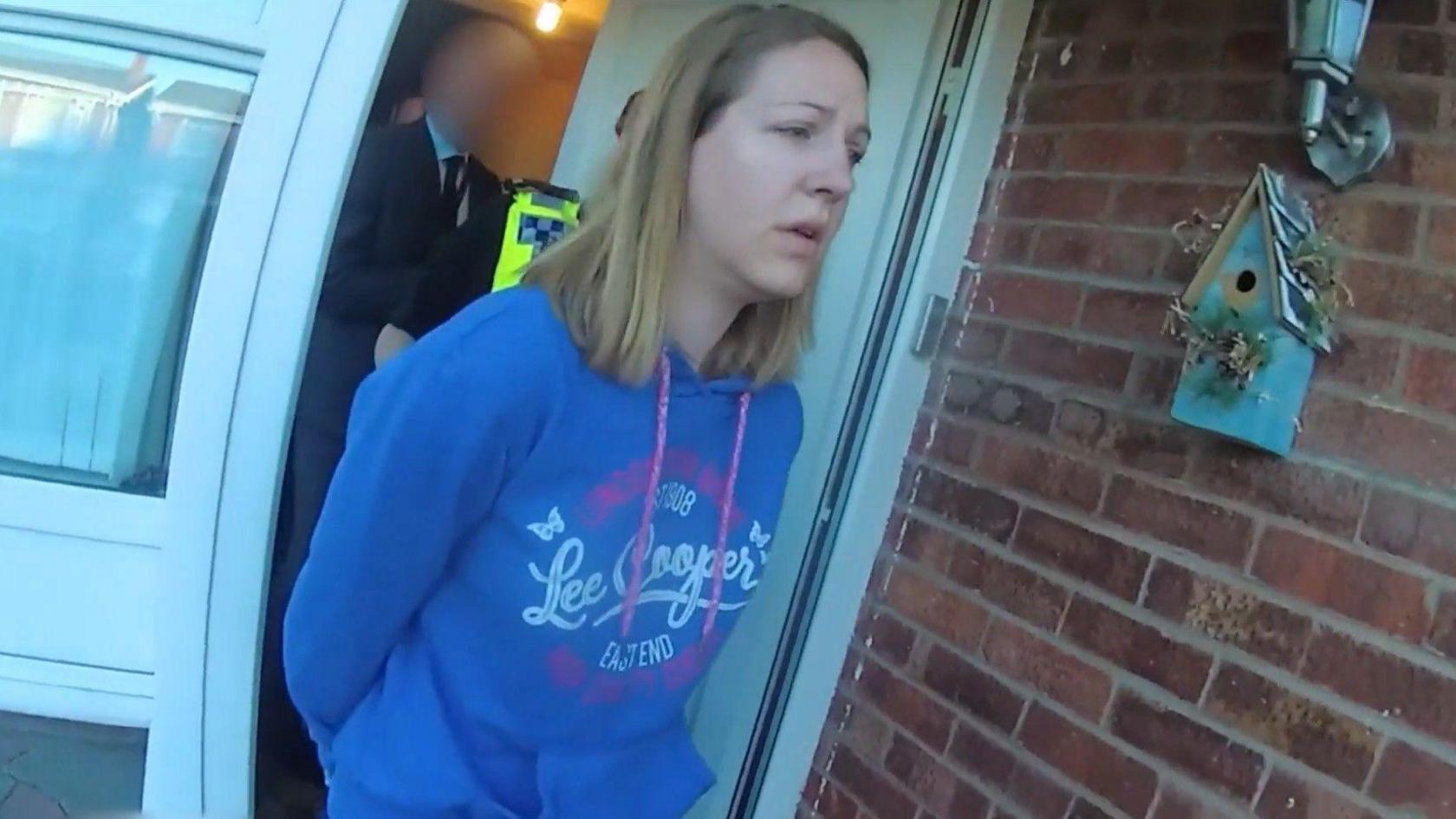 Lucy Letby wearing a blue hooded sweater coming out of her house in handcuffs with a police officer following her. She is looking past the camera, and is wearing her blonde/brown shoulder length hair down.