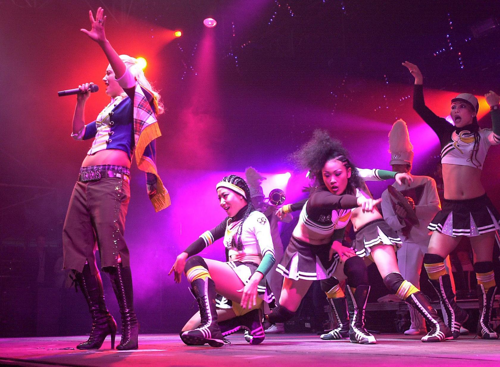 Gwen Stefani sings as a group of dancers in white and brown cheerleader outfits danced behind her. Gwen has long blond hair and is wearing boots, brown trousers and a blue, white and gold jacket