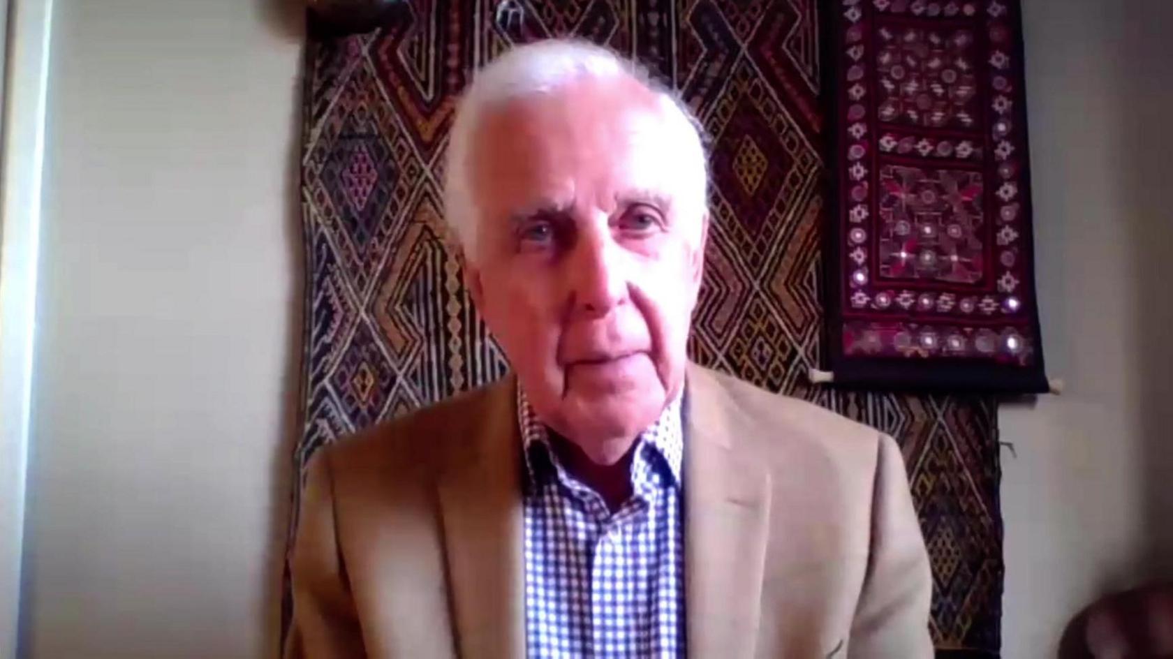 A still photo from a recorded webcam interview of a man with short white hair who is sat down and wearing a brown blazer and blue and white chequered shirt. A tapestry is hanging on a wall in the background which is red and black.