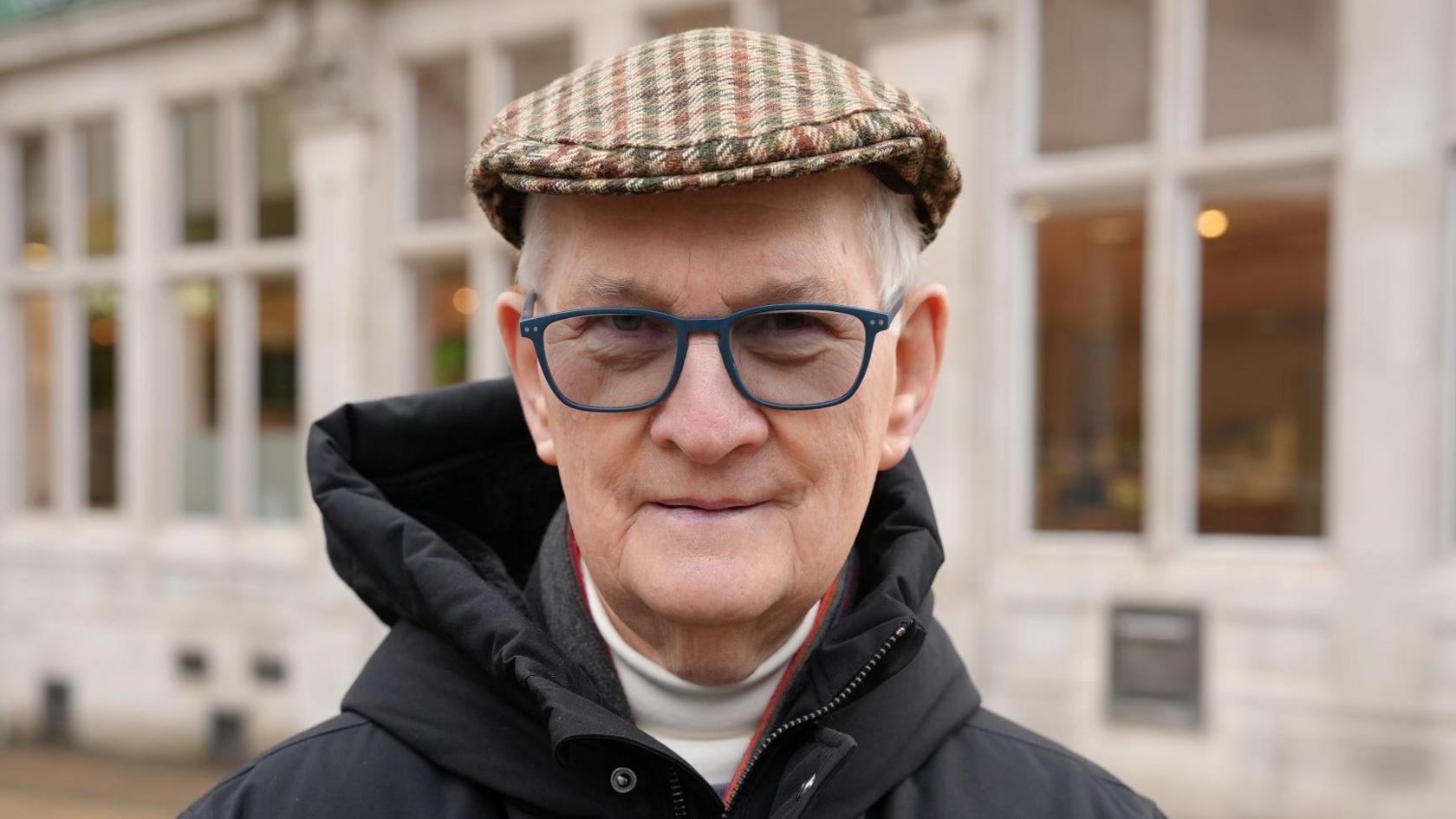 Robert Cato, who is wearing a brown, green and red striped cap. He has navy rimmed glasses on and is looking at the camera while wearing a black hooded coat. Behind him is a white building with many windows.