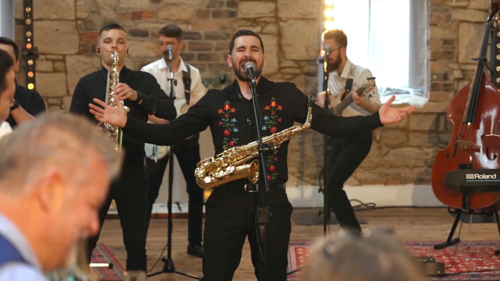 The band is playing at a wedding in large hall with stone walls. The lead singer is belting out a song while people look on.