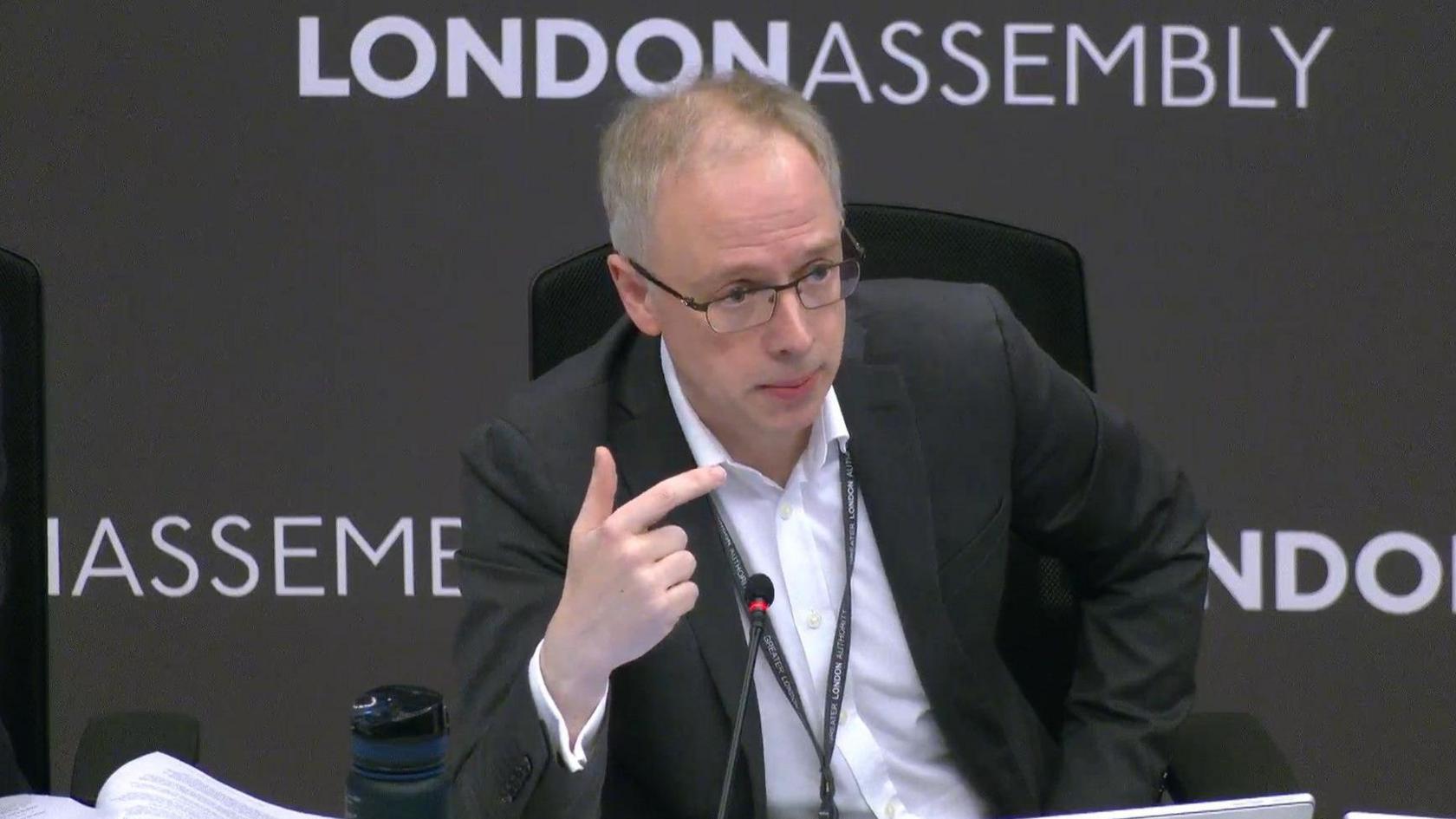 Richard Watts sits in front of a backdrop that says London Assembly. He is wearing a dark suit jacket and a white open-necked shirt, as well as a lanyard. There is a slim microphone on the table in front of him. 