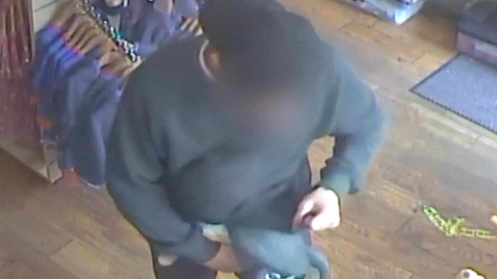 A screenshot from a video which shows a man dressed in a black jumper, blue jeans and a black cap, worn backwards, stuffing toys under his jumper. His face has been blurred.