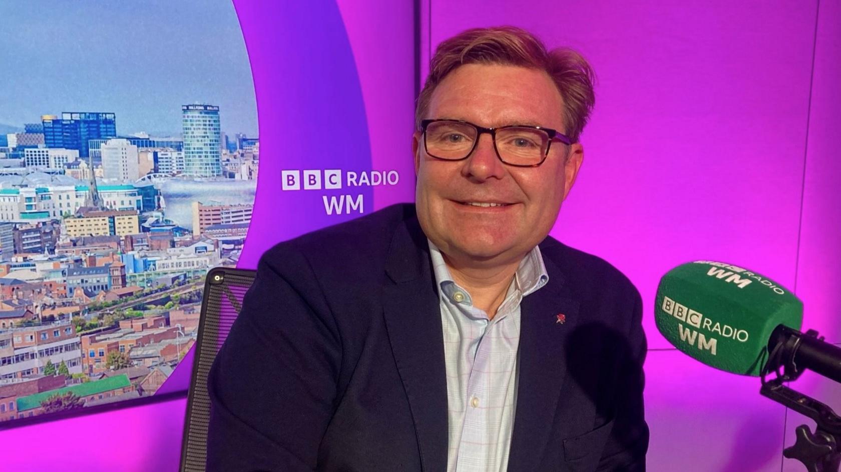 Birmingham City Council leader John Cotton sits in the BBC Radio WM studio in front of a green microphone and a purple backdrop of the Birmingham city centre skyline