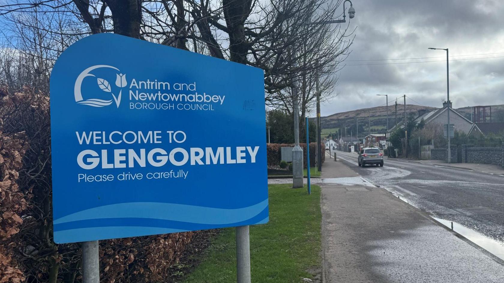 A large blue sign from Antrim and Newtownabbey Council is placed on the side of a two-way road. It reads 'Welcome to Glengormley, Please drive carefully' in white writing. There is a grey car on the road driving slightly in front of the sign.