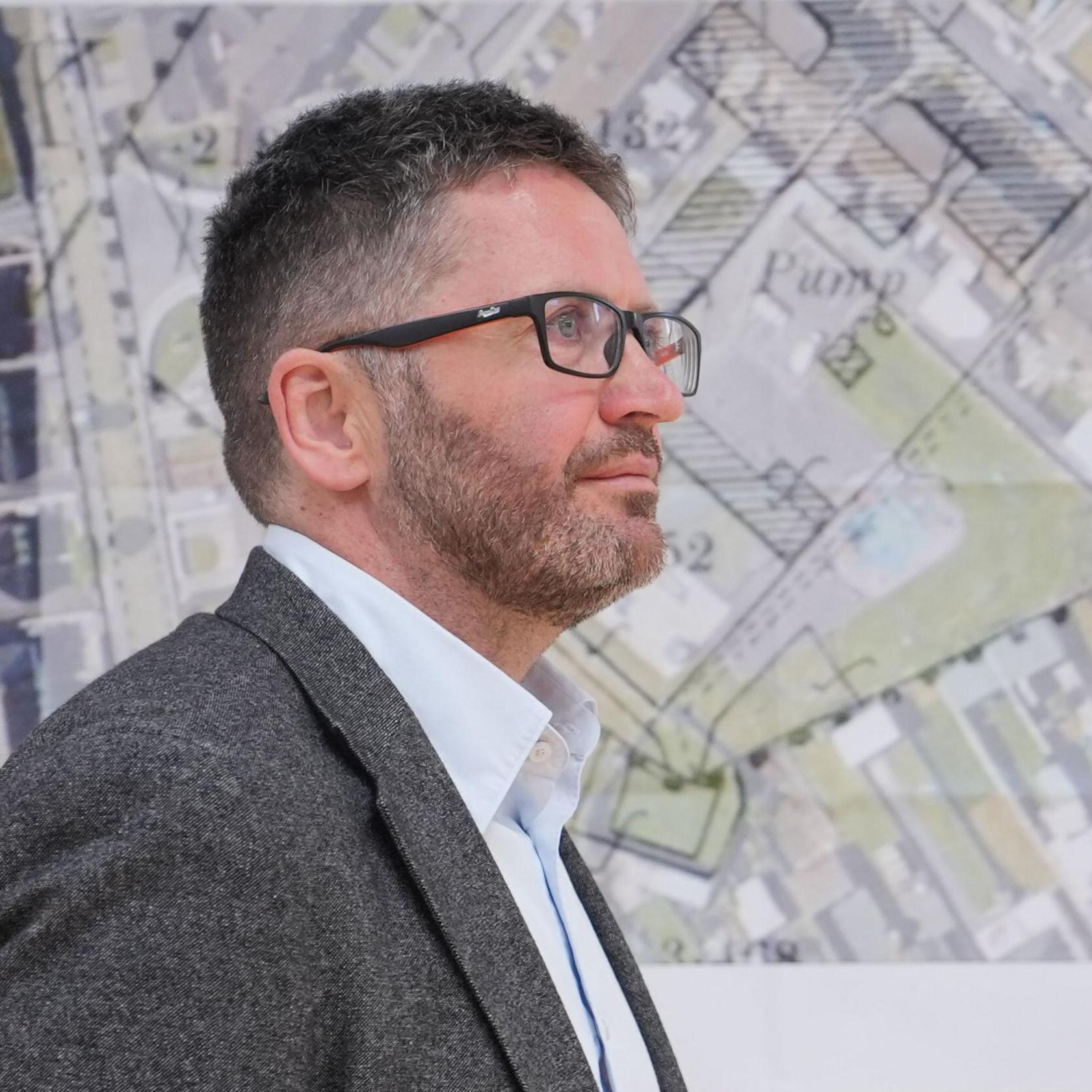 A man wearing a grey jacket, light-coloured shirt and glasses looks off to the side. He has short cropped dark hair with flecks of grey. He is standing in front of survey map which is overlaid on an image of a site