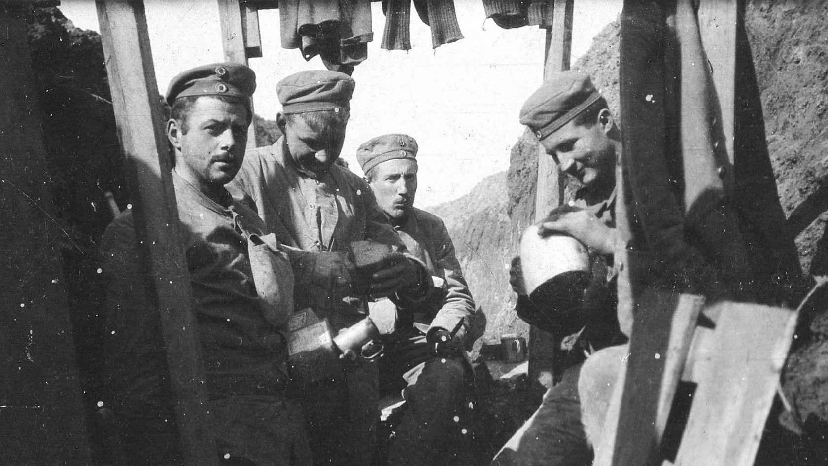 Four German soldiers sit in a trench holding what appear to be, canisters of water