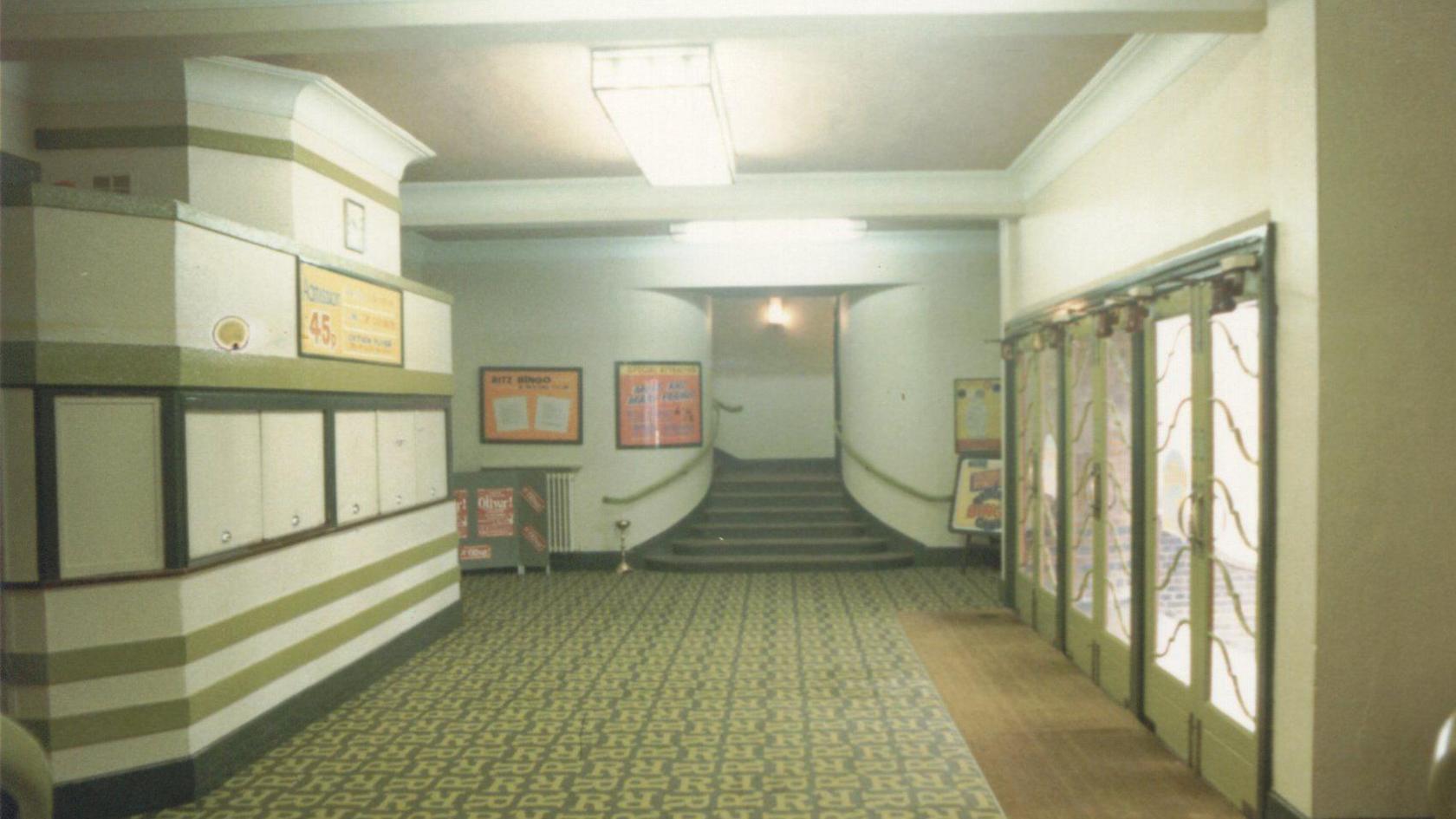 An inside of a building showing carpet, with R on it, a staircase, a close up foyer, with green stipes, large front doors, white walls and art deco lights in the ceiling.