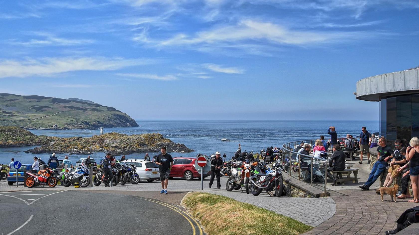 The Sound car park on a sunny day. It is busy with many bikes, cars and people. The Calf of Man is in the background with the sea in between, and the cafe is on the right.