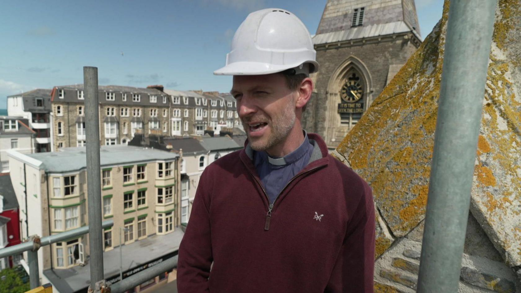 Pip and Jim's Church vicar Reverend Mark Ruoff stands on scaffolding at the venue. He has a burgundy jumper on over his tunic and dog collar. He is wearing a white hard hat.