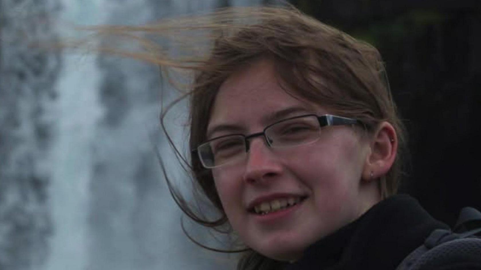 Headshot picture of a woman wearing glasses and brown hair who is smiling. Her hair is tied back but is windswept. She is stood outside and is wearing a black coat.