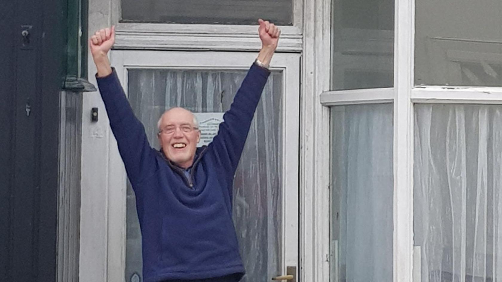 Charlie Lambert celebrating outside Norman Nicholson's house after the property was bought in February. Mr Lambert is smiling and his hands are up in the air. he is wearing a navy blue sweatshirt and has glasses. The house has large front windows with white panes. 