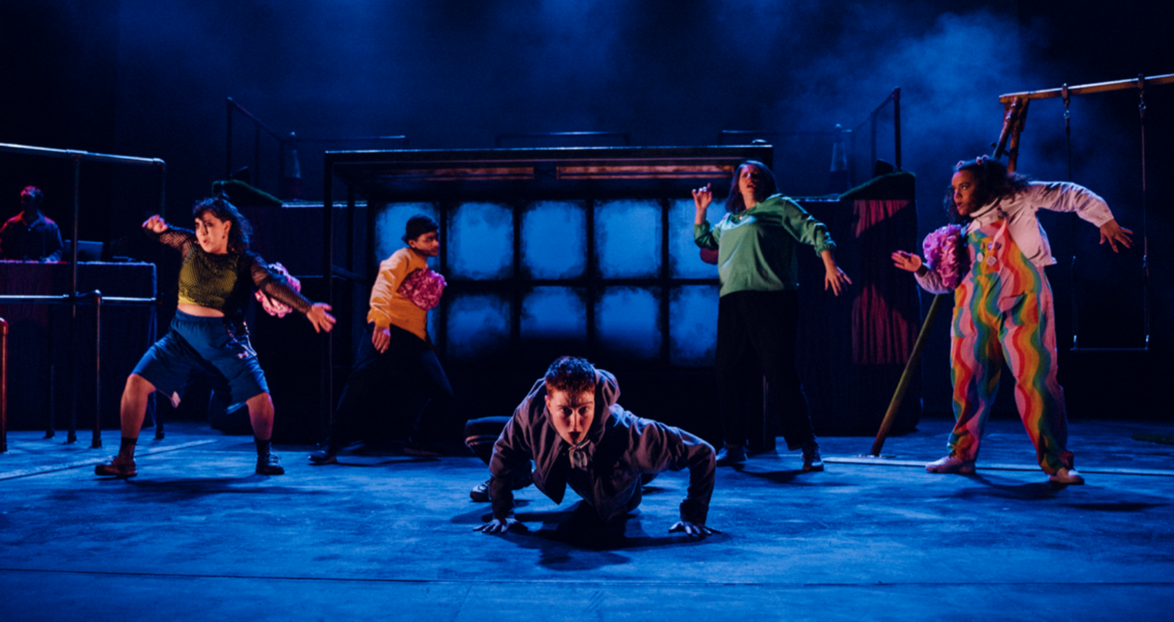 Five performers captured under dark blue stage lights. The centre performer is crawling across the floor and stares directly at the camera while other characters pose in abstract positions around them. 