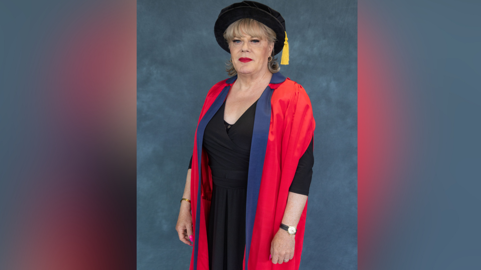 Suzy Izzard receiving her honorary doctorate from the University of Sussex. She is wearing a black dress and a red graduation gown with purple piping. Suzy is wearing a black mortarboard.