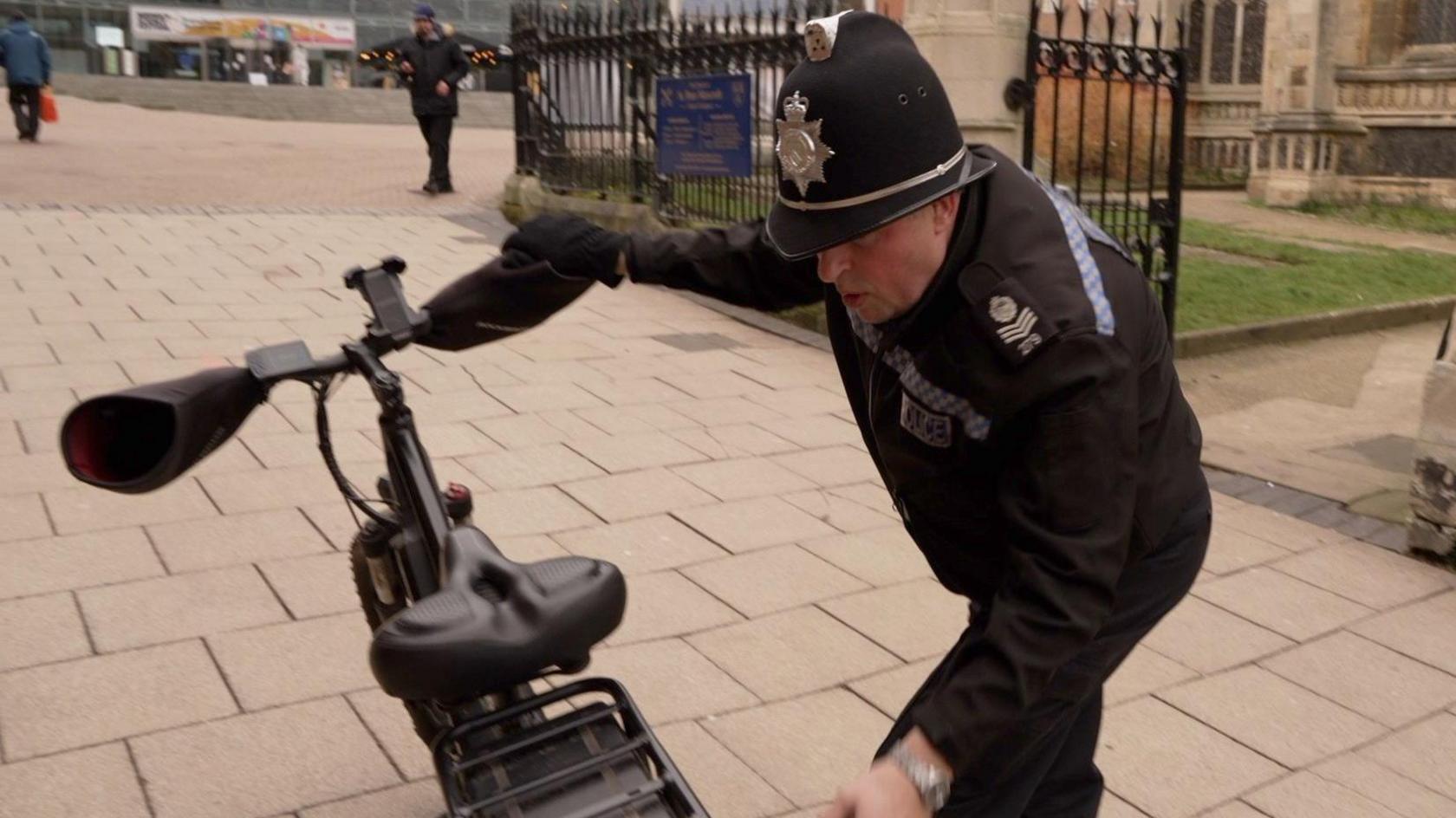 A police officer is looking at a black bike. He is stood next to the bike and is wearing a black police uniform and hat.