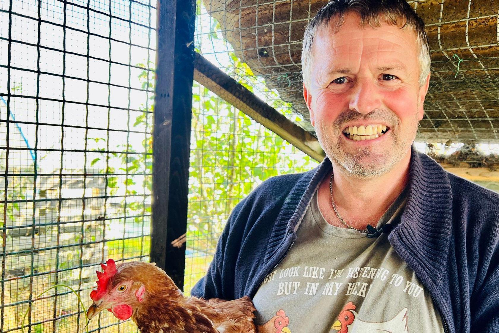 A man holds a hen in a coop on an allotment in Hessle. He is smiling and wearing a blue cardigan and a grey T-shirt.