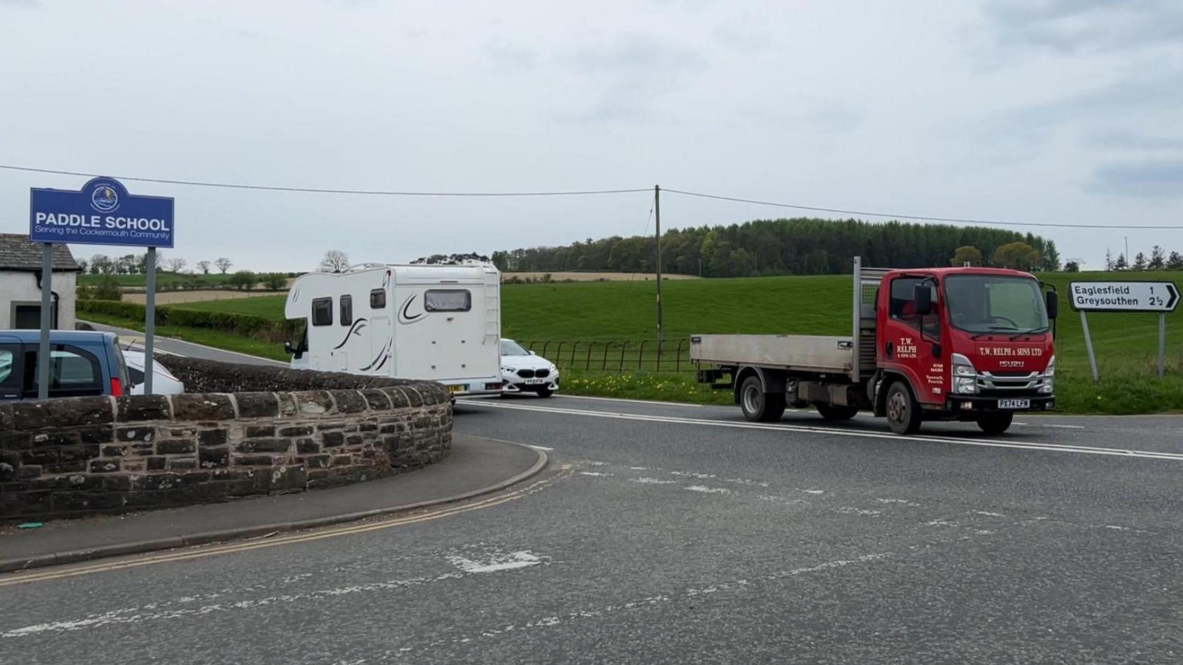 A crossroads with traffic travelling along the main road. A blue sign for "Paddle School" is visible on the left, inside the school grounds' stone boundary wall, which runs along the corner of the junction.