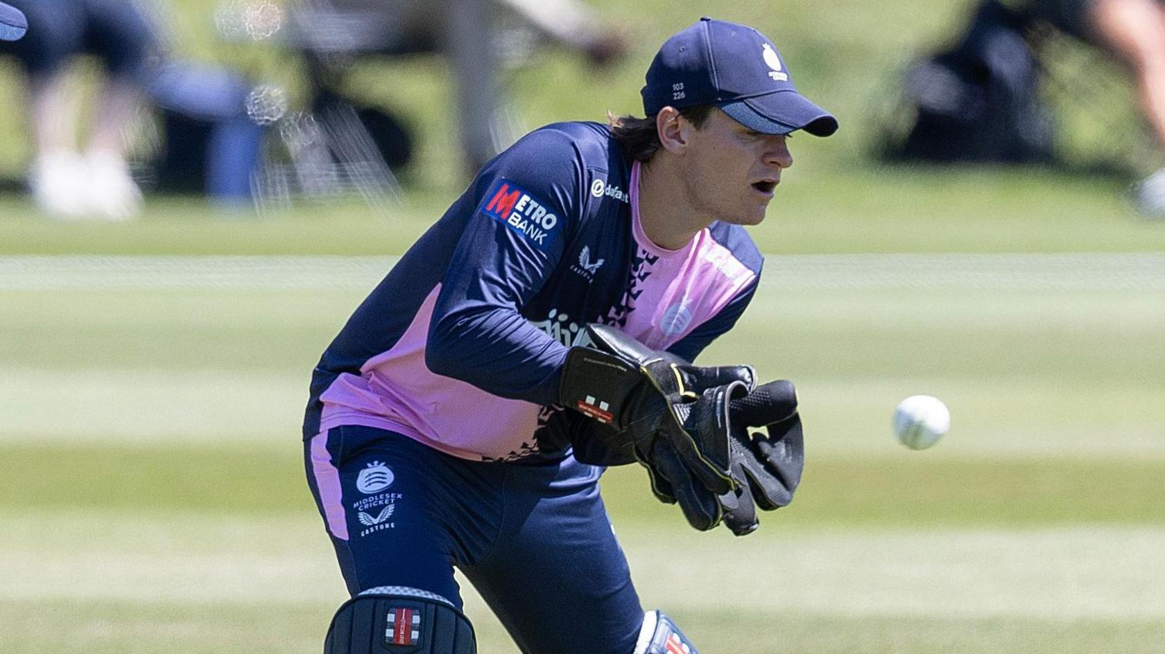 Jack Davies keeping wicket in gloves, cap and pads, with navy and pink shirt and navy trousers