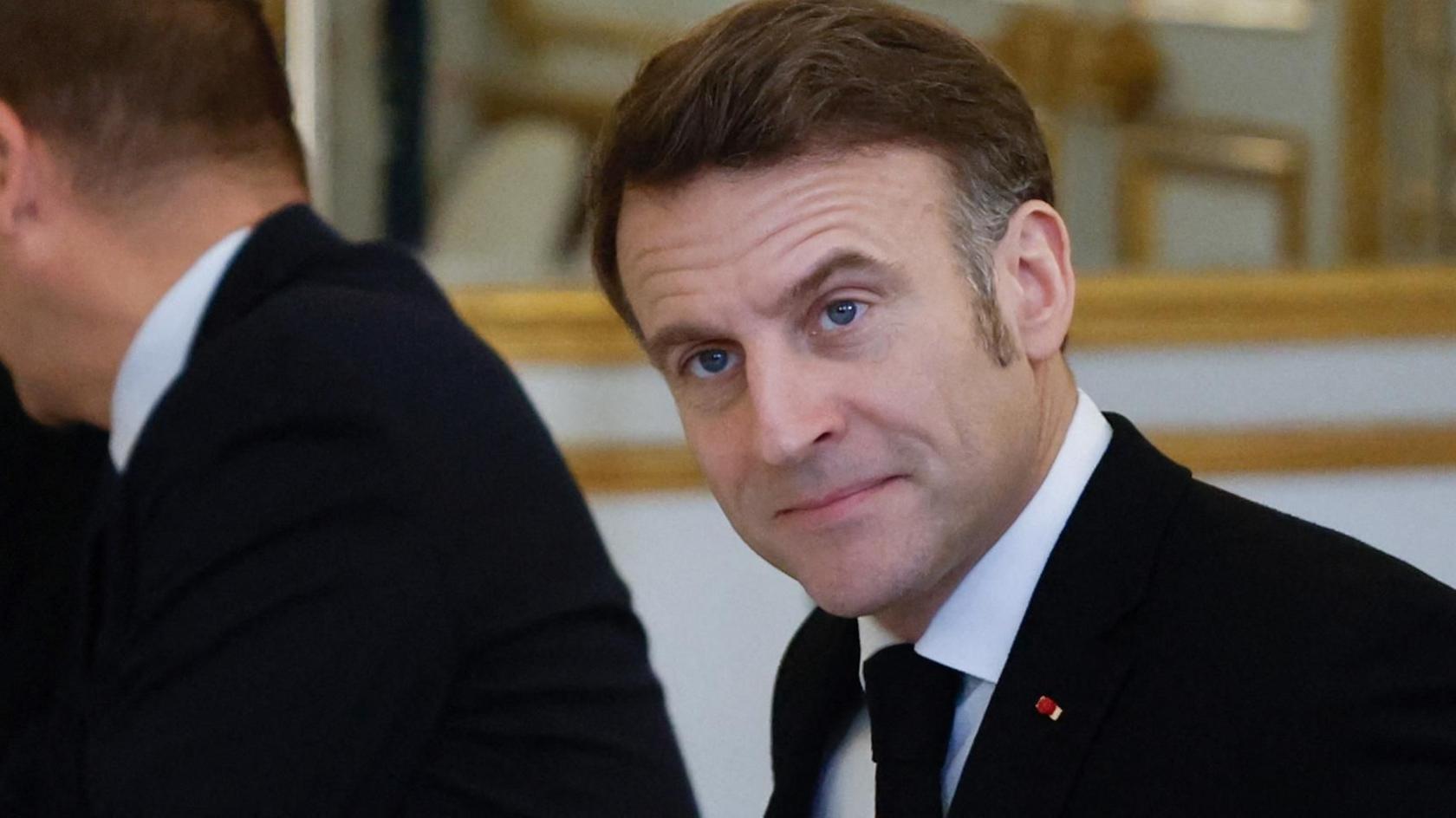 Emmanuel Macron in a black suit and tie looking off camera with a serious face.