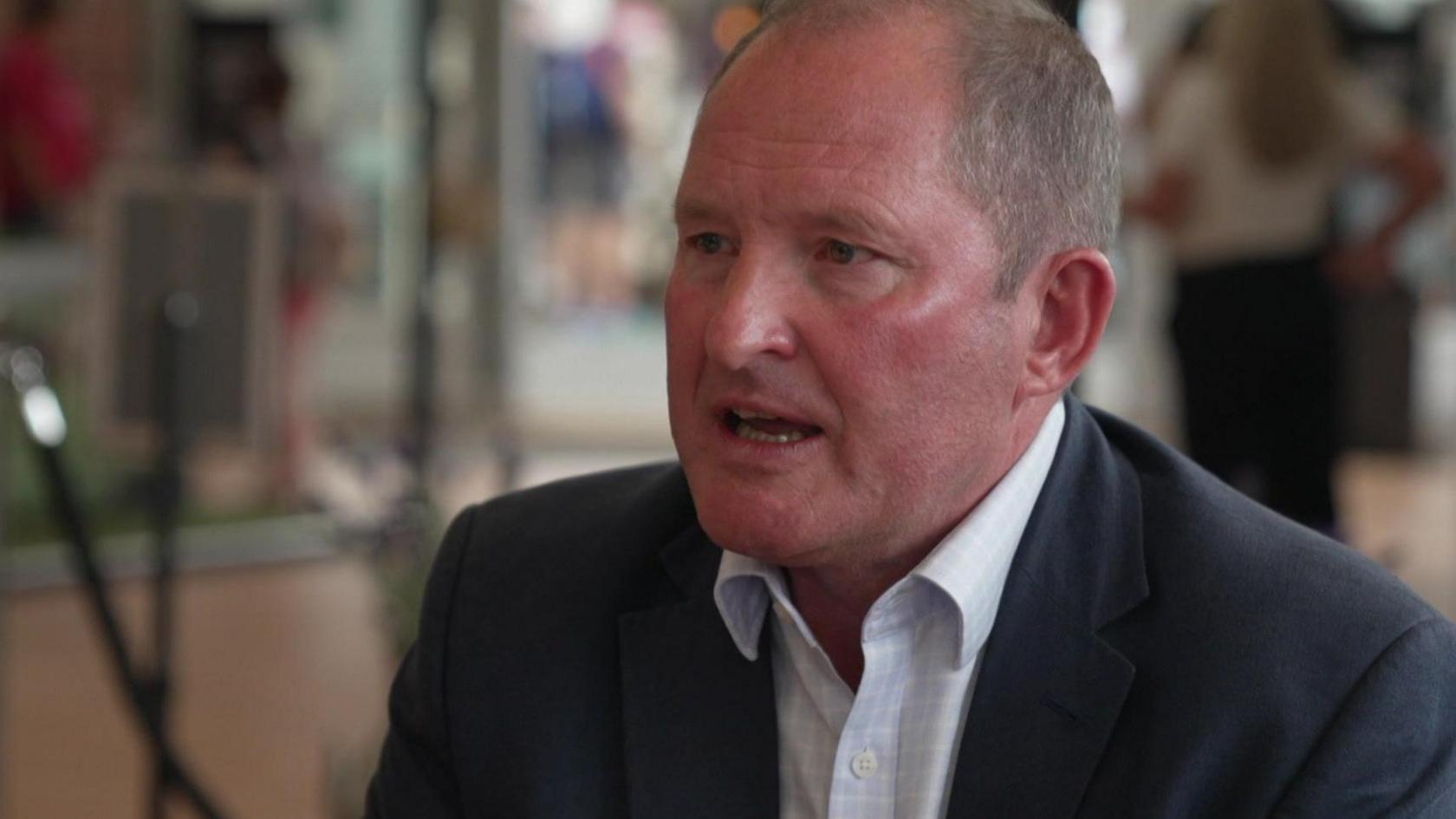 Steve McLean being interviewed with a blurred background behind him and wearing a dark suit and light shirt open at the neck.