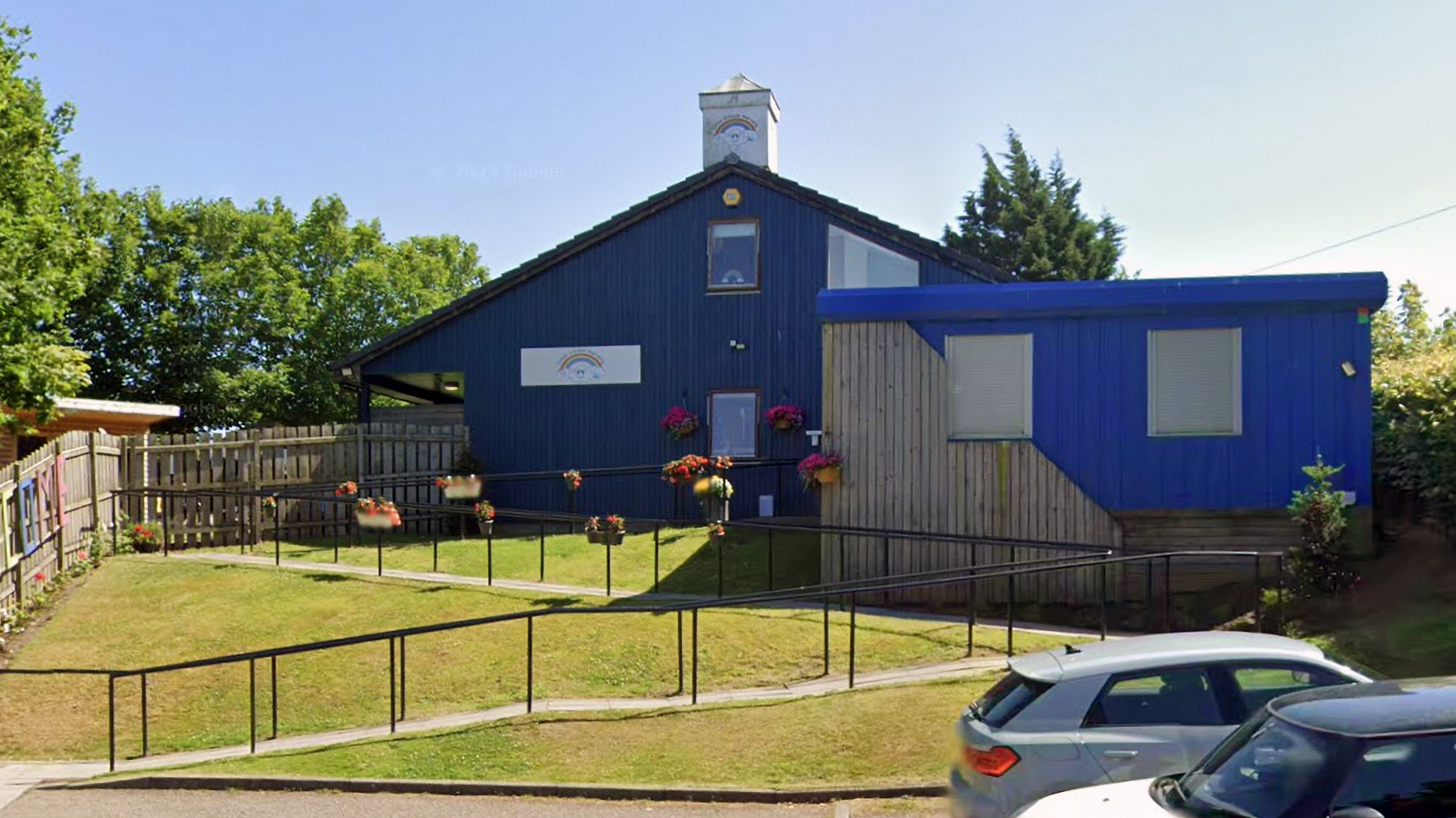Single-story building with dark blue and light blue exterior walls, a sloped roof, and a small white cupola on top. The front area has a ramp with black railings leading to the entrance, and several hanging flower baskets add colour along the ramp. Two cars are parked in the foreground, and the background includes green trees and a clear blue sky
