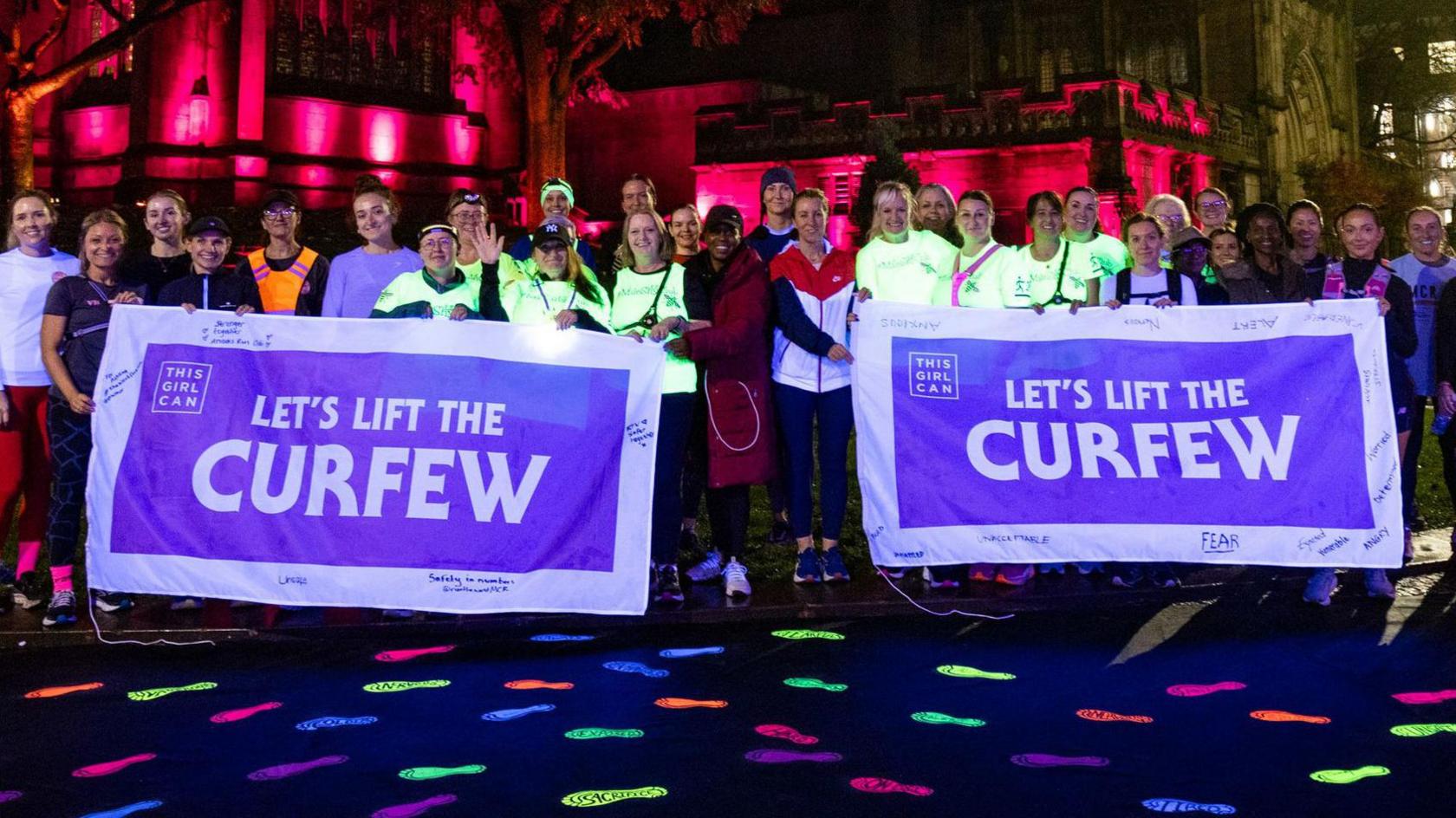 A group of people dressed in exercise clothing. They are holding big purple signs saying "Let's lift the curfew". There are glow-in-the-dark footprints on the floor. 