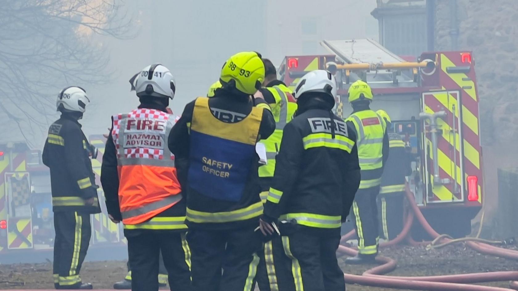 Firefighters, a safety officer and hazmat advisor stand in a group wearing high-vis firefighting attire and hard hats. The air around them is smoke-filled and multiple fire engines can be seen in the background.