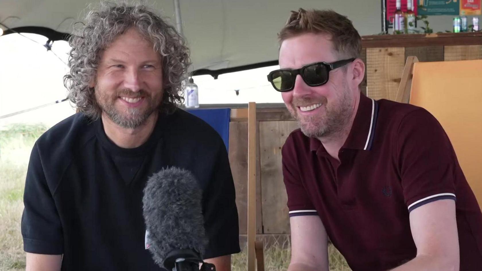 A wide shot of Simon Rix and Ricky Wilson being interviewed. Simon has long dark curly hair and is wearing a black top. Ricky has short dark hair and is wearing a maroon polo shirt, and sunglasses.